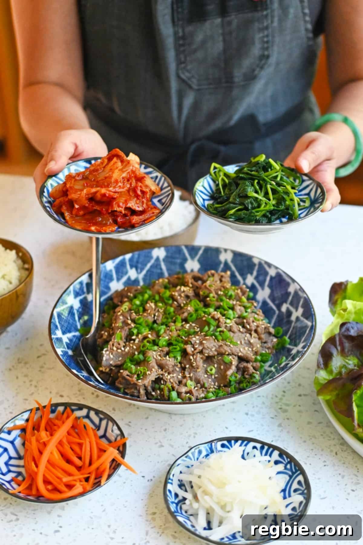 Two hands holding small bowls of kimchi and cooked spinach above a bowl of bulgogi and other small plates of banchan.