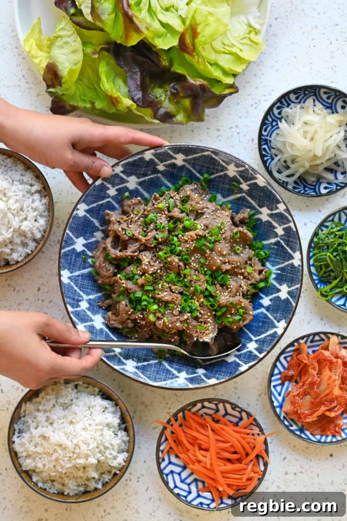 A hand is placing a serving spoon into a bowl of paleo bulgogi. There are small bowls of vegetables and steamed rice surrounding the beef.