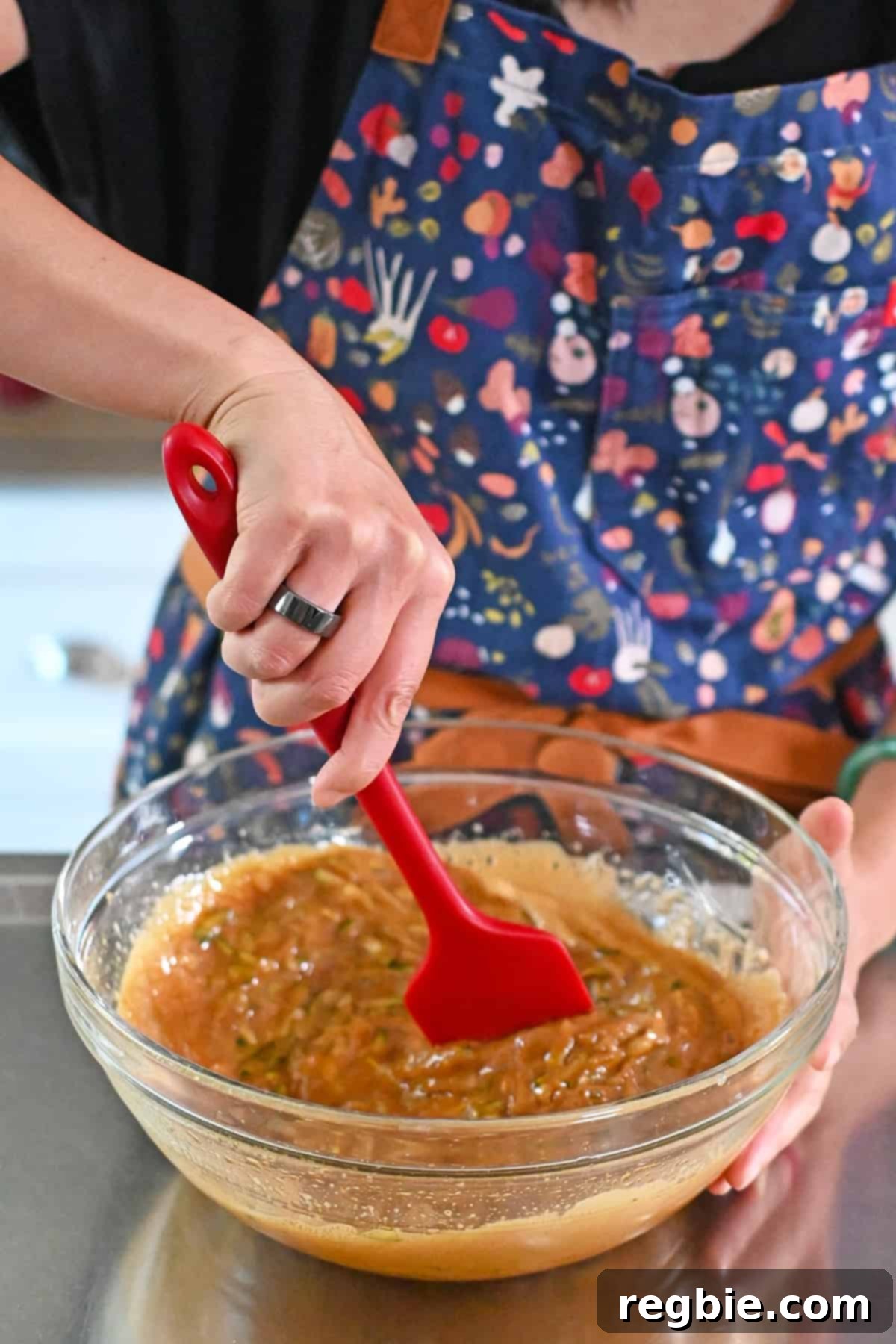 Stirring the shredded zucchini into the coconut sugar and egg mixture with a red silicone spatula, ensuring even distribution.