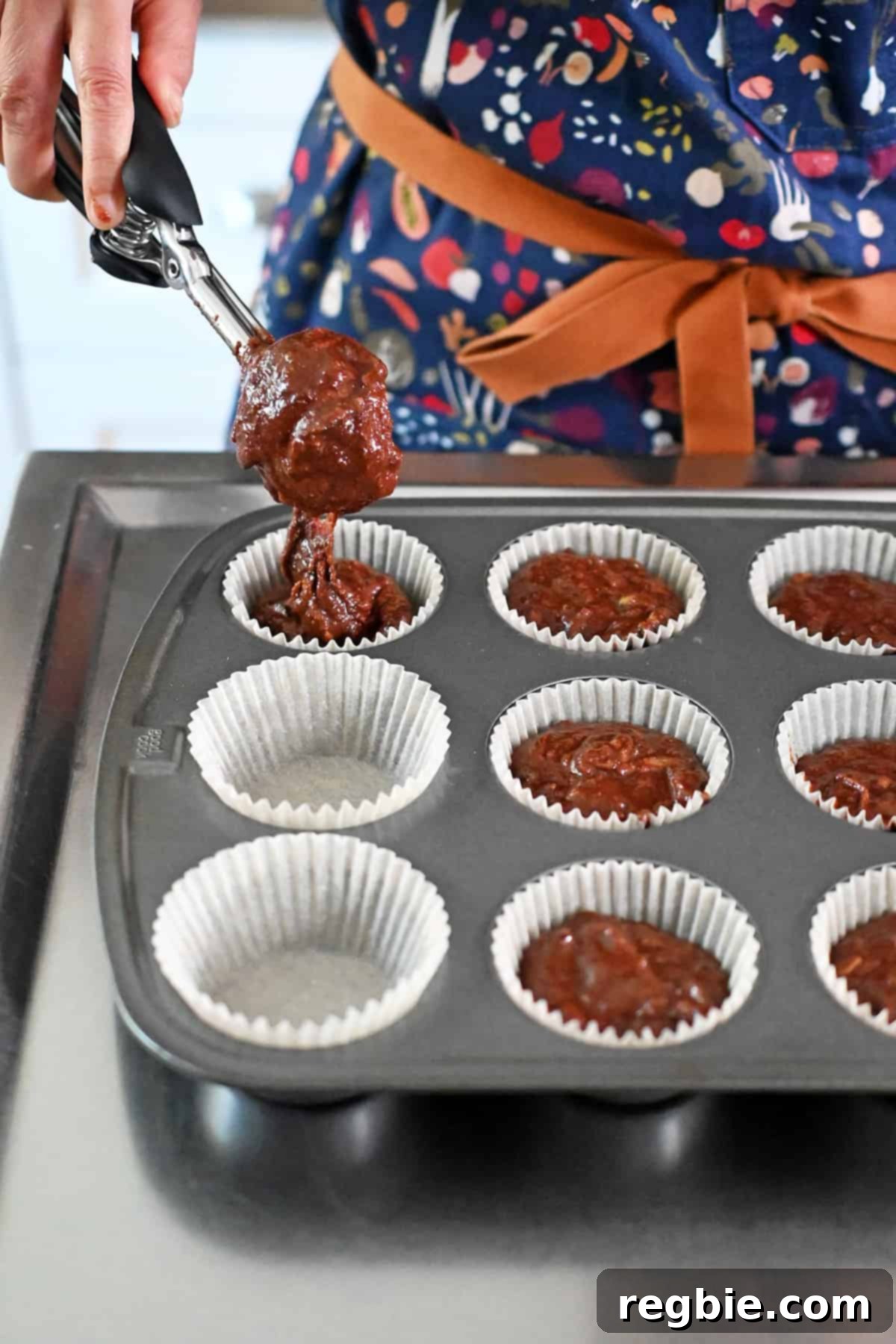 Using an ice cream scooper to transfer the chocolate zucchini muffin batter into a parchment paper lined muffin tin for baking.
