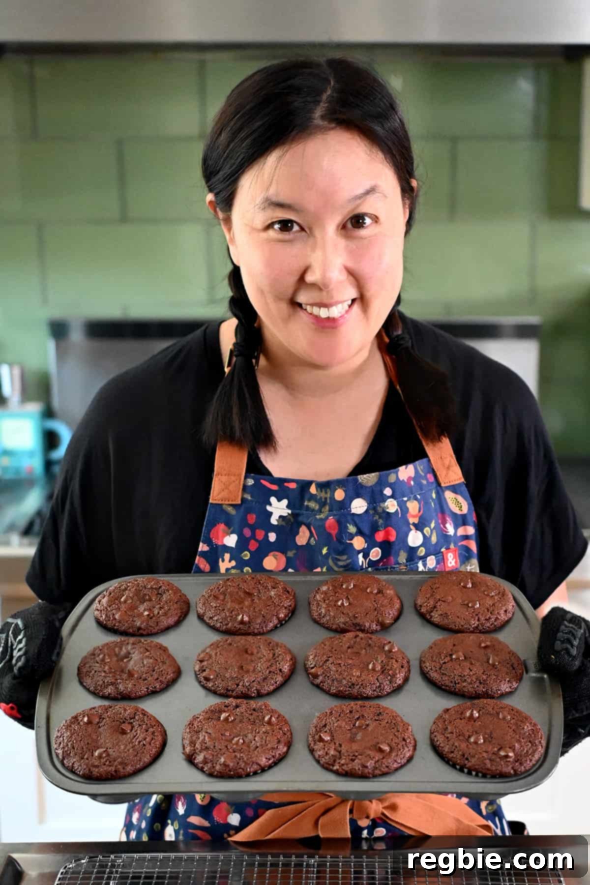 A smiling Asian woman in braids is holding up a muffin pan filled with freshly baked paleo chocolate zucchini muffins.