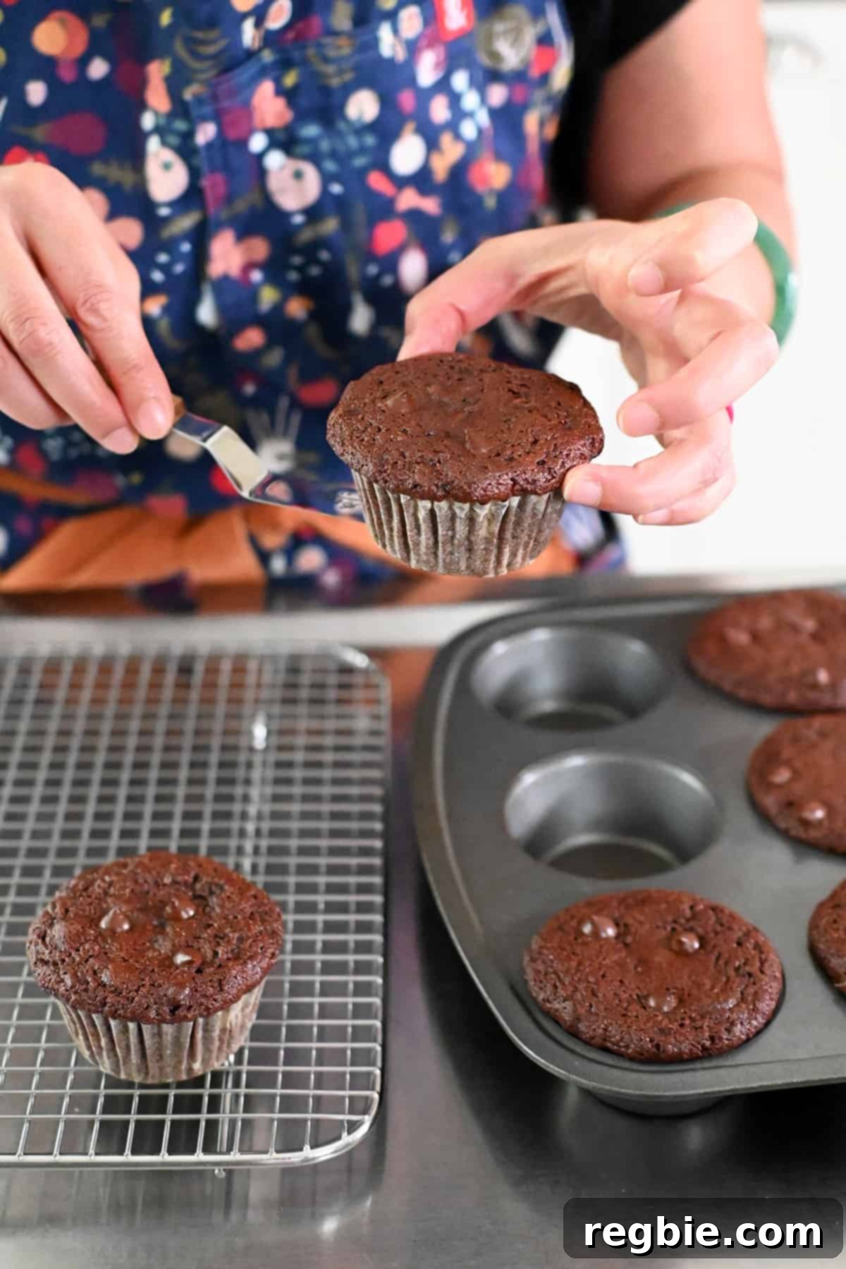 Lifting a freshly baked chocolate zucchini muffin from the tin and putting it on a cooling rack to cool down.