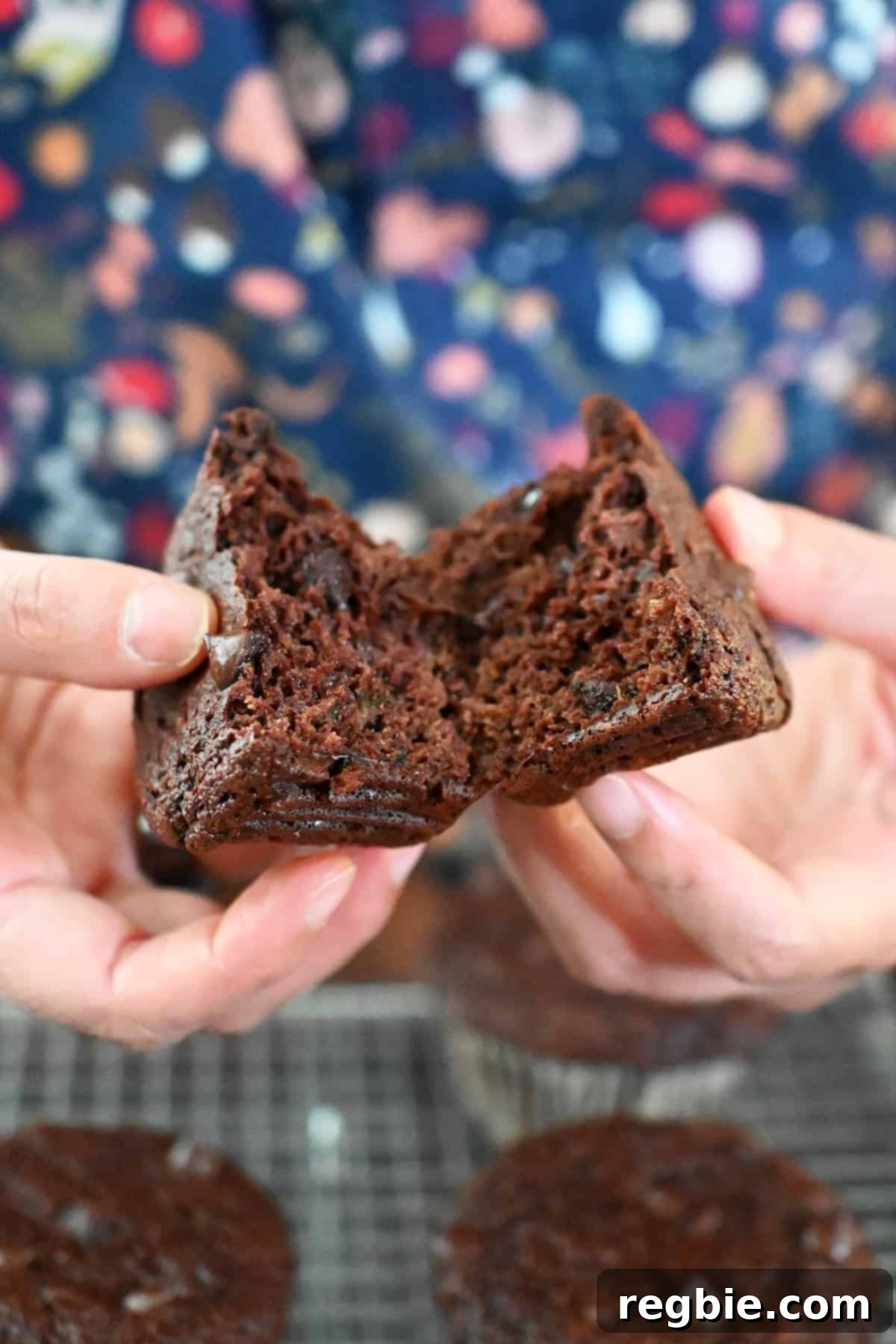 Two hands are breaking a chocolate zucchini muffin in half, revealing the moist, chocolatey interior with no visible zucchini pieces.