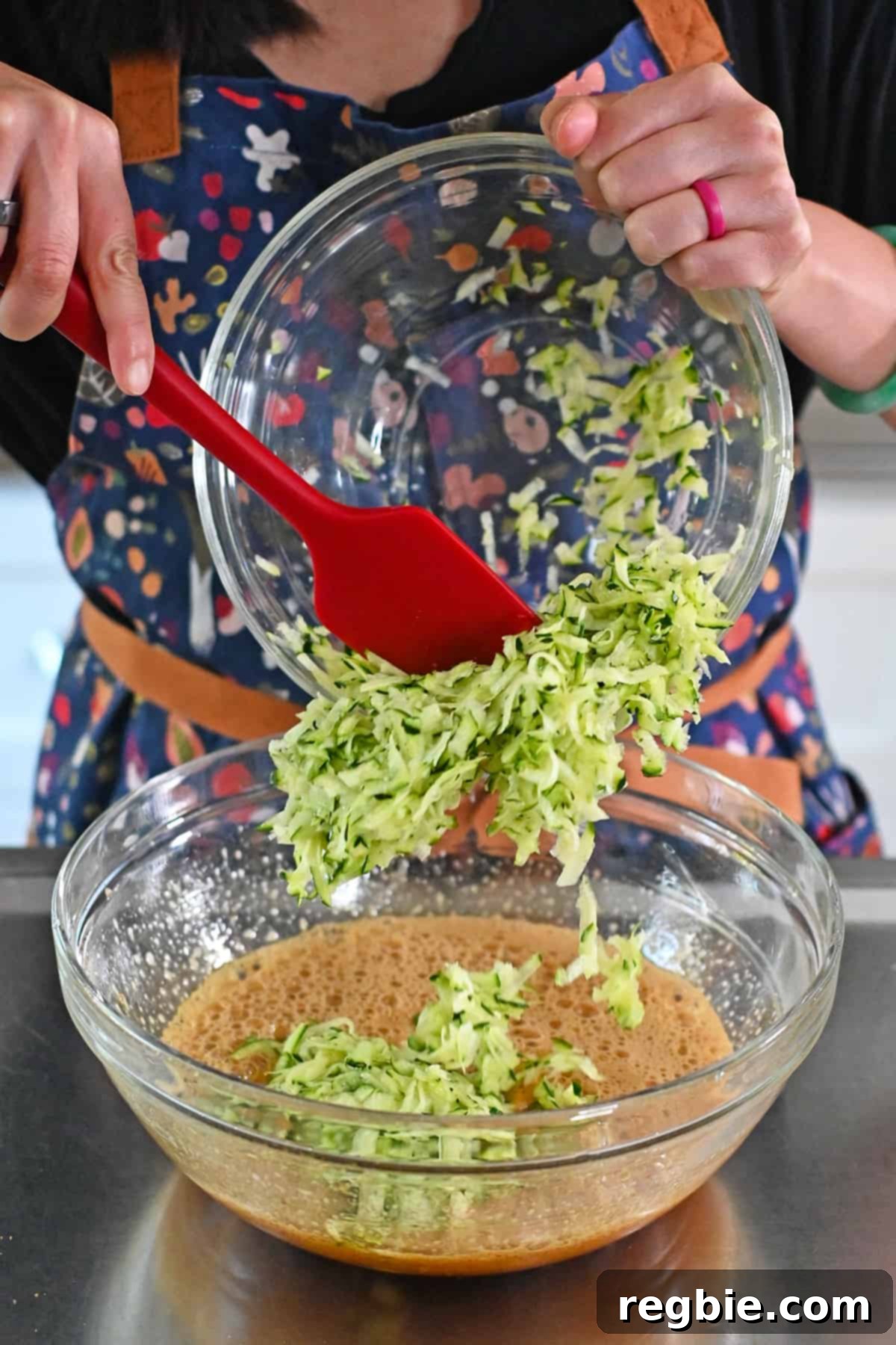 Adding shredded zucchini the bowl with the wet ingredients to make chocolate zucchini muffins, preparing for the next step.