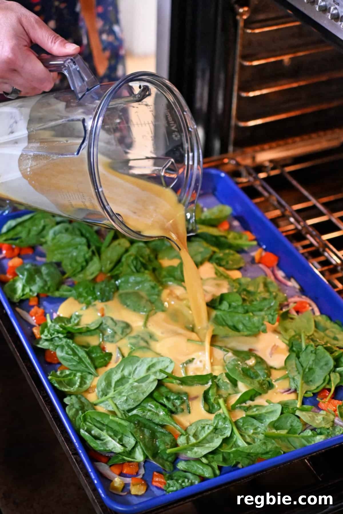 Pouring blended raw eggs from a Vitamix blender cup into a blue rimmed sheet pan filled with baby spinach, roasted sliced red onions, and roasted red bell peppers.