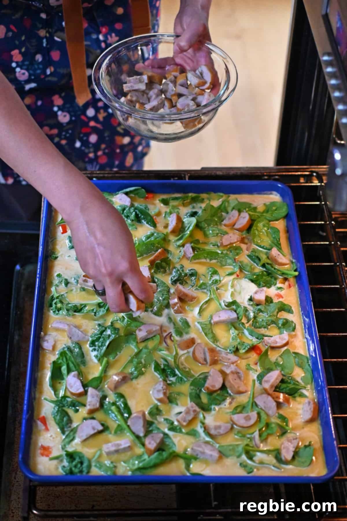 A hand is placing thinly sliced chicken apple sausage onto an unbaked blue sheet pan filled with raw scrambled eggs and vegetables in an open oven.