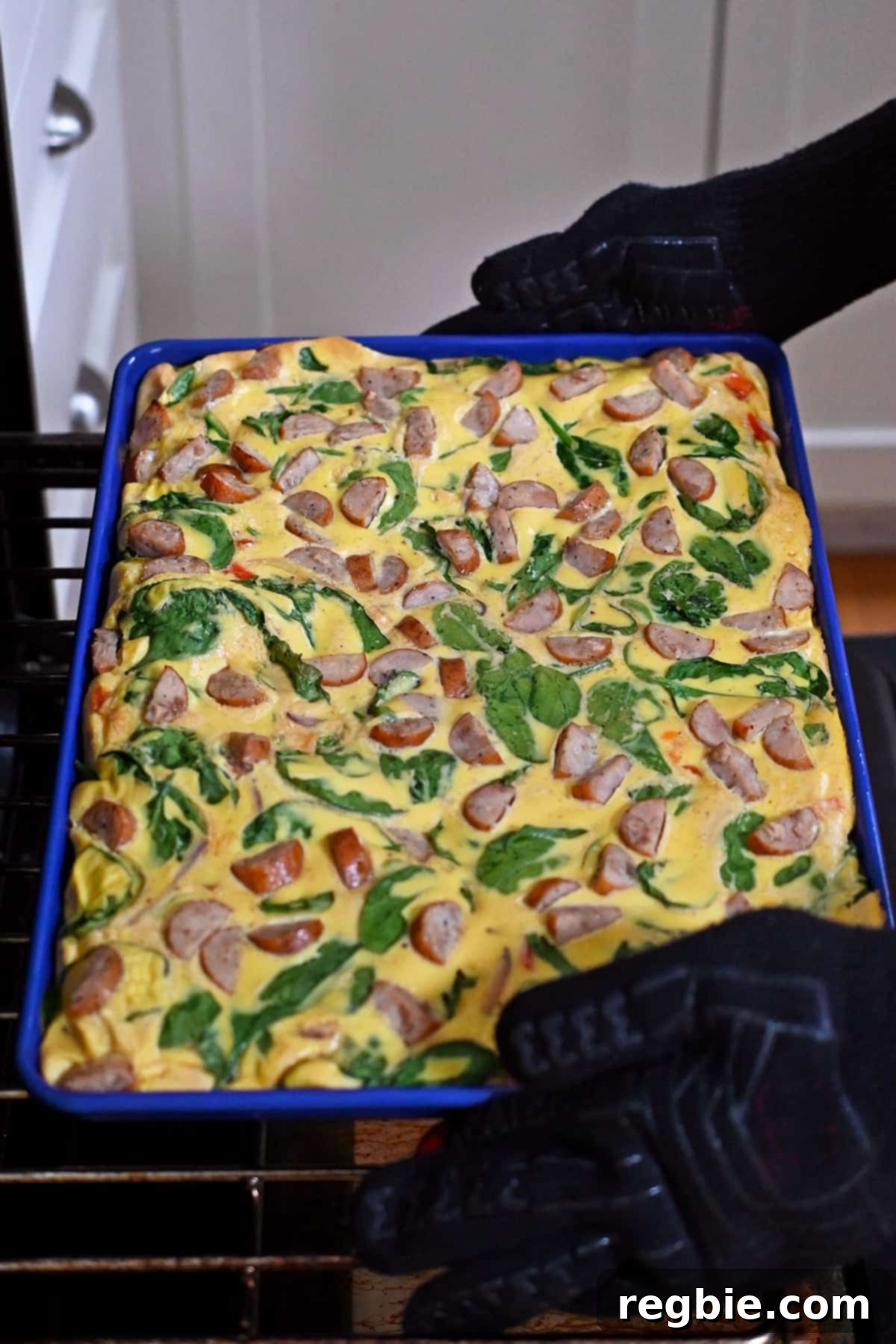 Removing a freshly baked sheet pan filled with scrambled eggs, veggies, and sliced chicken apple sausage out of an oven.