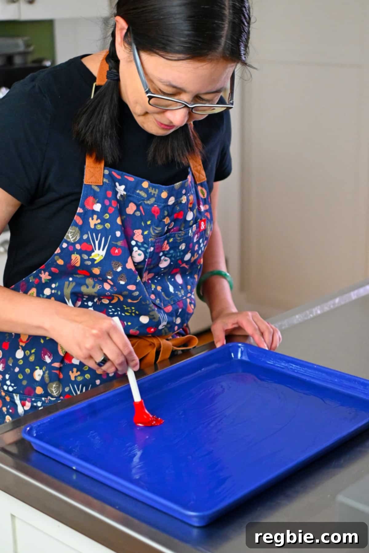 A smiling Asian woman is brushing oil on a blue rimmed baking sheet to make sheet pan eggs.