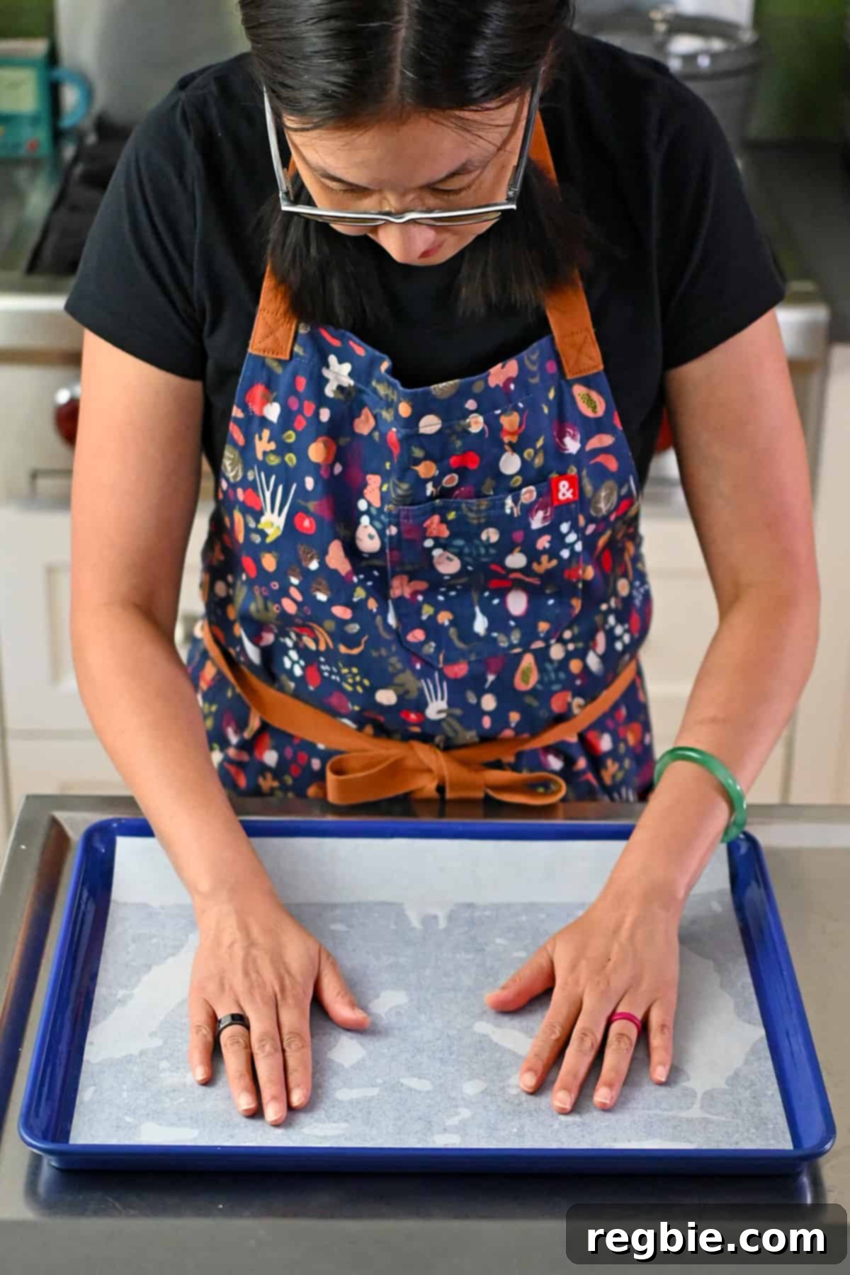 An Asian woman is placing a piece of parchment paper into a greased blue rimmed sheet pan,