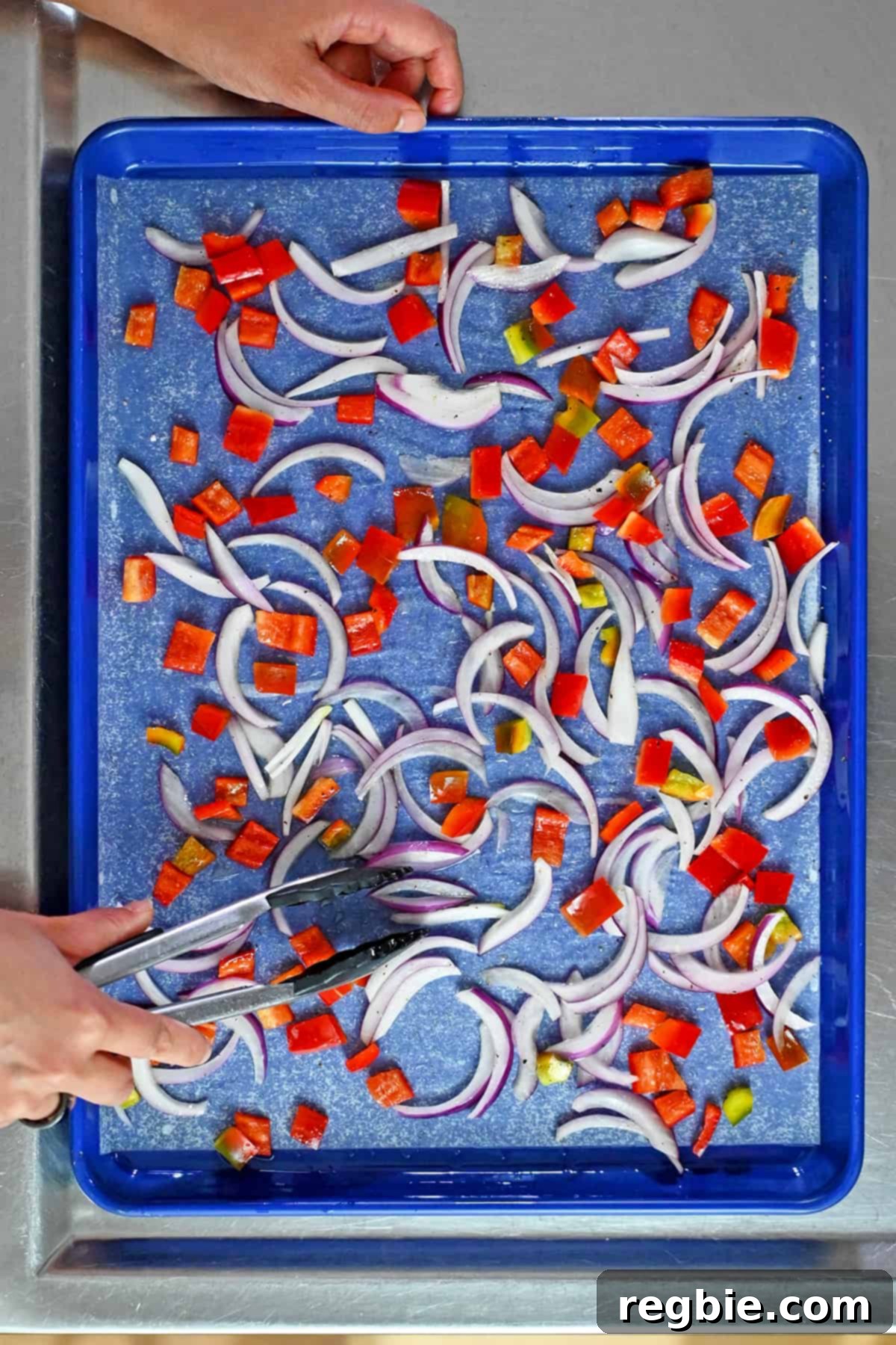 A pair of tongs is rearranging thinly sliced red onions and diced bell peppers in a single layer on a parchment lined rimmed baking sheet.
