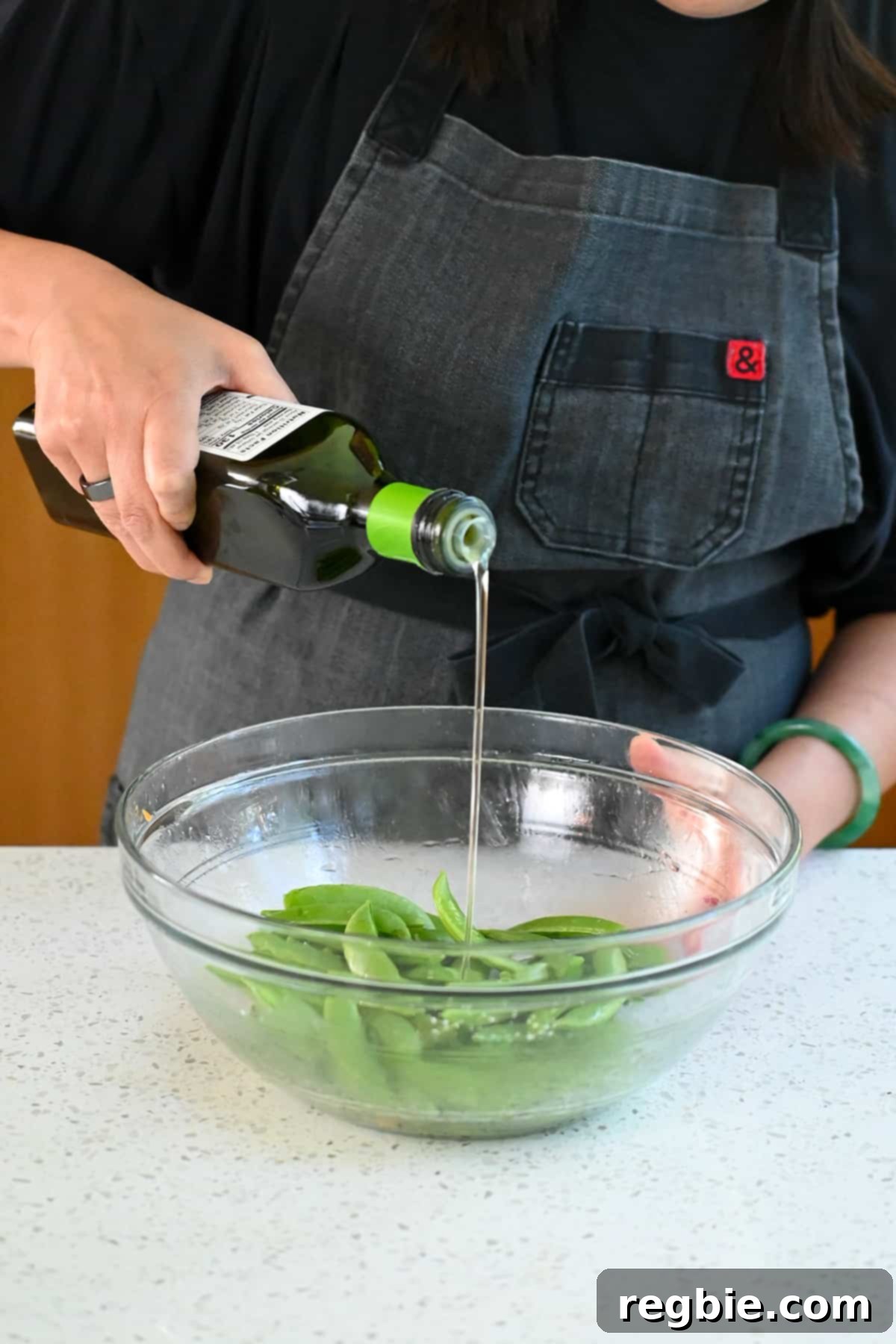 Sheet Pan Cashew Chicken: Paleo & Whole30 Perfection 11 Pouring a stream of avocado oil from a bottle into a bowl already filled with vibrant raw sugar snap peas, ready for tossing.