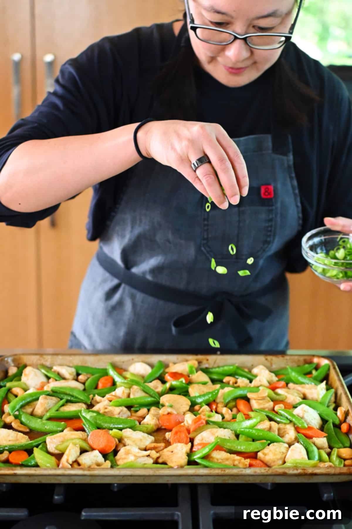 Sheet Pan Cashew Chicken: Paleo & Whole30 Perfection 18 An Asian woman gracefully sprinkling thinly sliced green onions on top of a generous tray of Sheet Pan Cashew Chicken, adding a fresh, vibrant garnish.