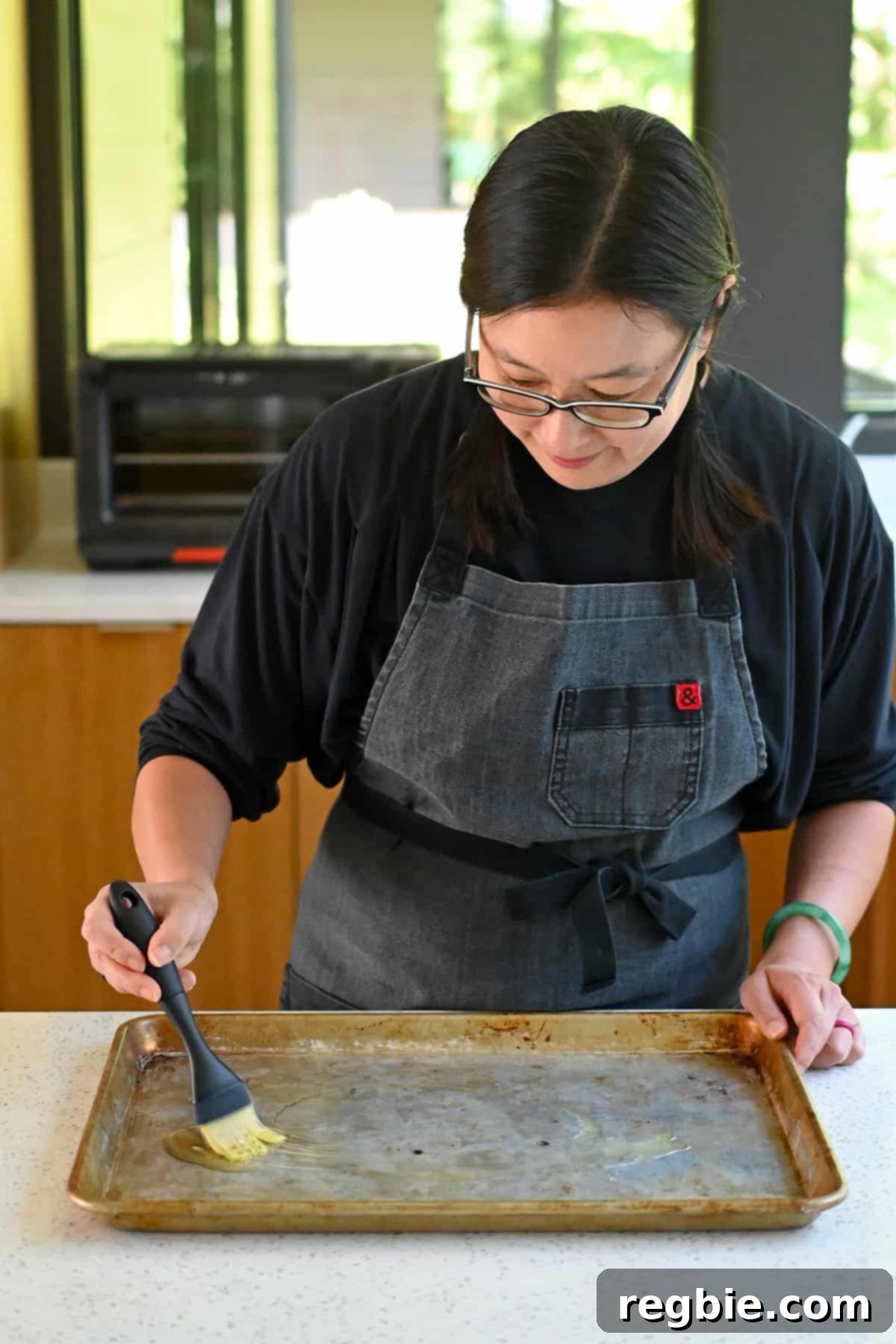 Sheet Pan Cashew Chicken: Paleo & Whole30 Perfection 5 An Asian woman in glasses is brushing avocado oil on a rimmed baking sheet, preparing it for the sheet pan cashew chicken.