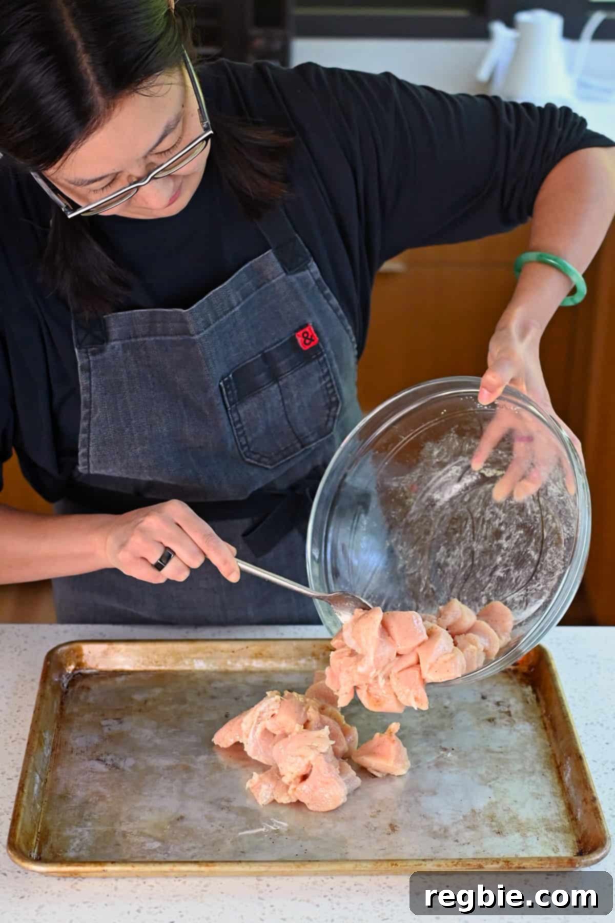 Sheet Pan Cashew Chicken: Paleo & Whole30 Perfection 7 An Asian woman carefully placing seasoned raw chicken breast cubes onto a greased rimmed baking sheet, ready for roasting.