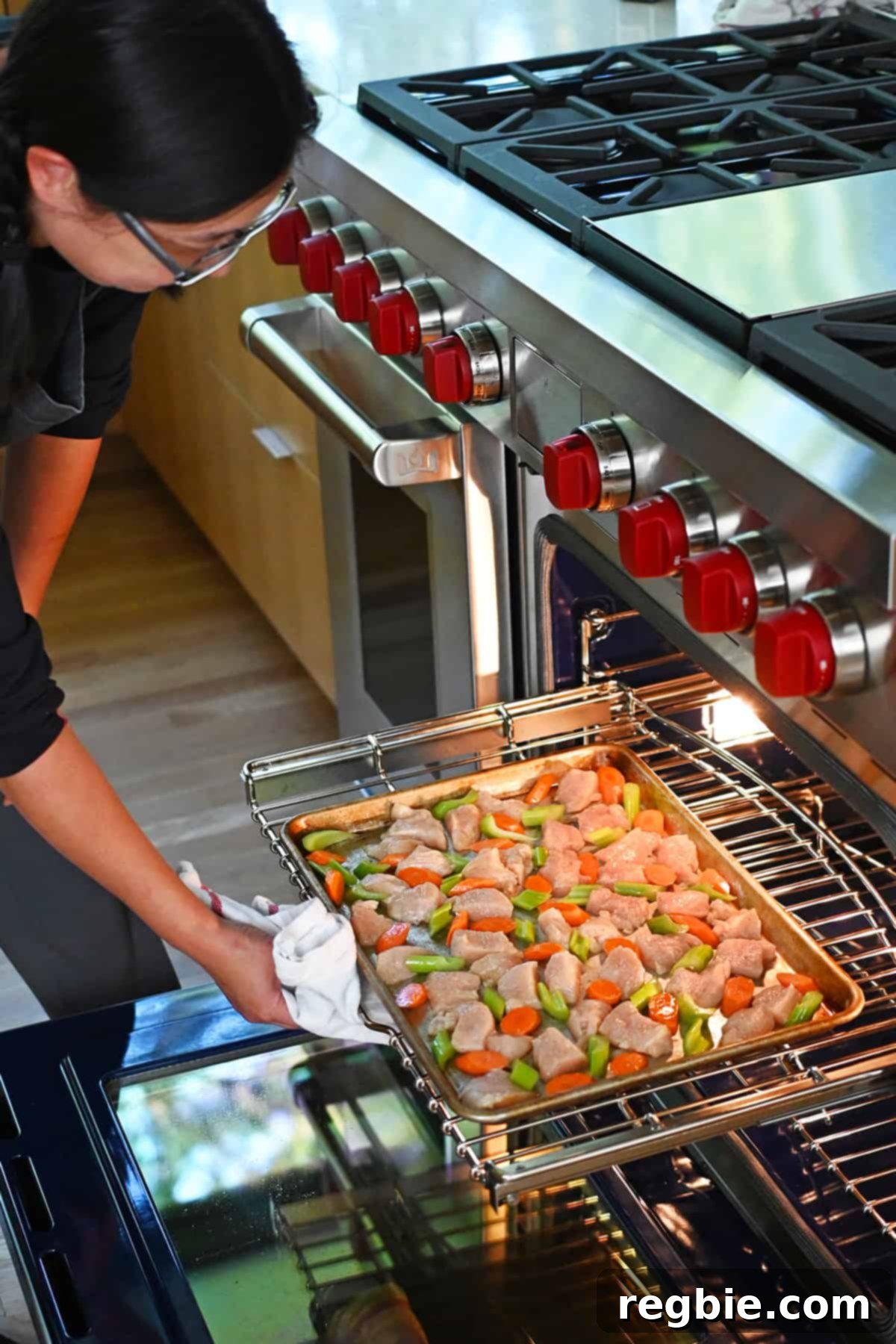 Sheet Pan Cashew Chicken: Paleo & Whole30 Perfection 10 An Asian woman gently sliding a sheet pan, laden with raw chicken breast cubes, sliced carrots, and sliced celery, into a hot, open oven.