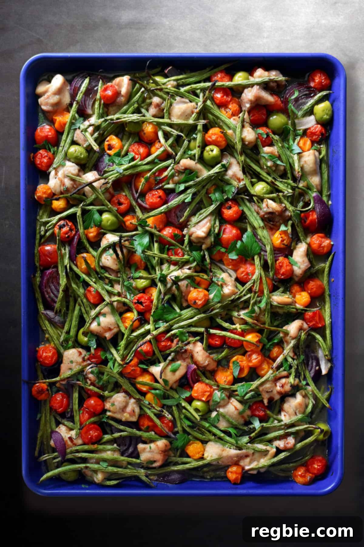 An overhead shot of a blue sheet pan filled with cooked cubed chicken thighs, roasted green beans, roasted cherry tomatoes, red onion wedges, green olives, fresh herbs, and a drizzle of balsamic vinegar, ready to serve.
