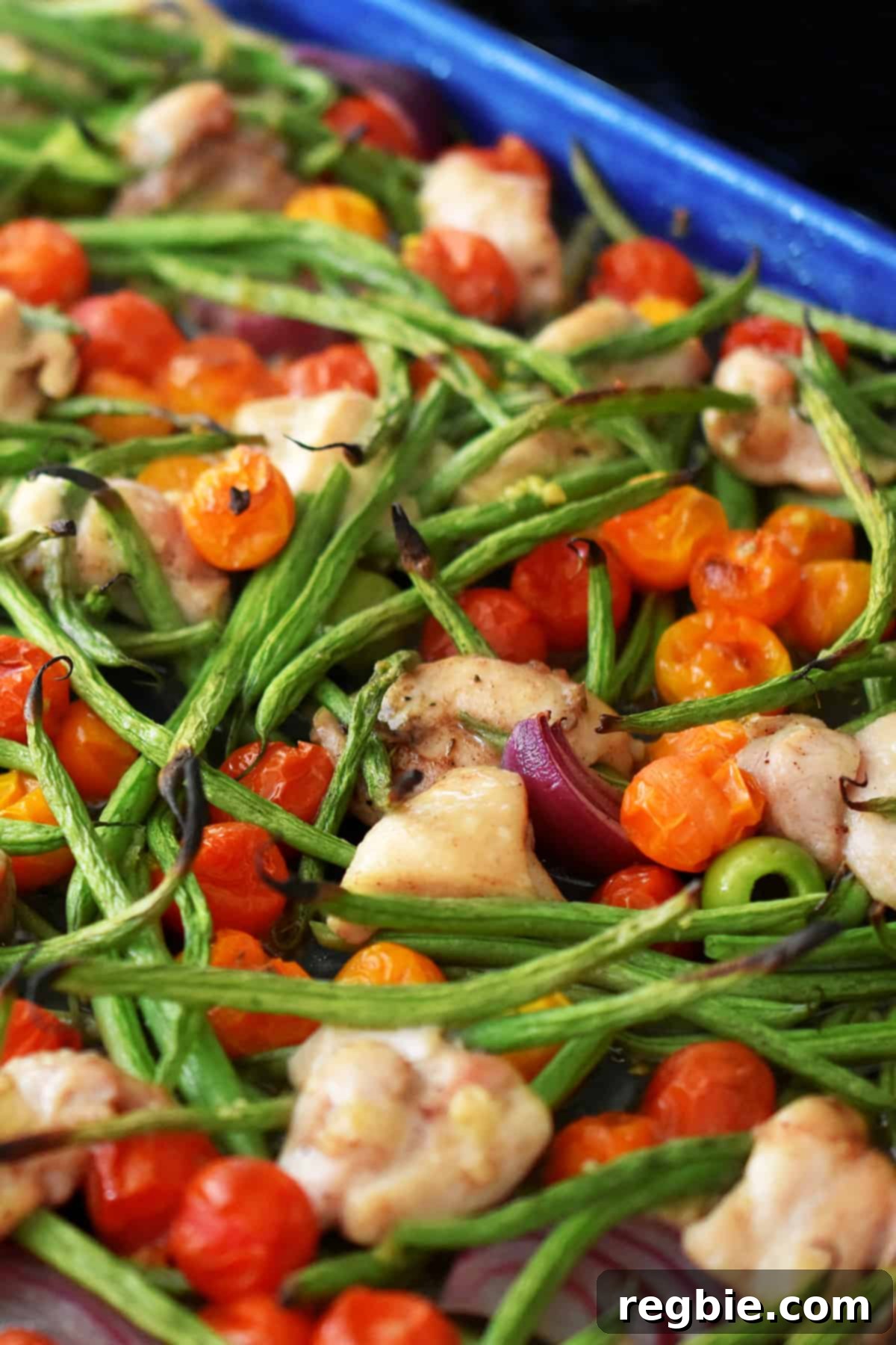 A close-up shot of the finished sheet pan Italian chicken dinner, fresh out of the oven, highlighting charred green beans, wrinkled cherry tomatoes, and perfectly cooked chicken thigh pieces.