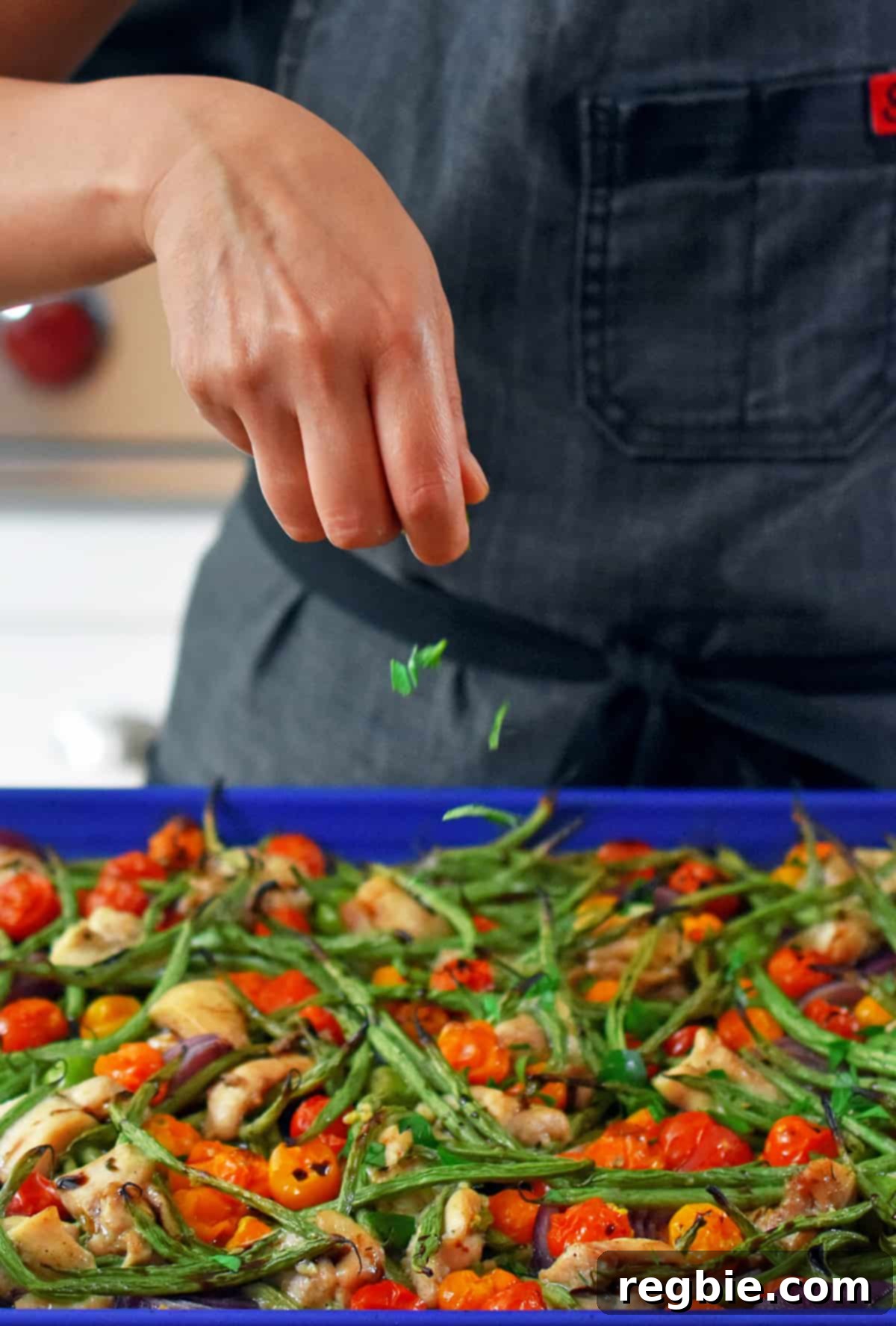 A hand adding minced fresh herbs, likely parsley or basil, as a garnish to the sheet pan Italian chicken dinner directly after it has come out of the oven, enhancing its visual appeal and fresh flavor.