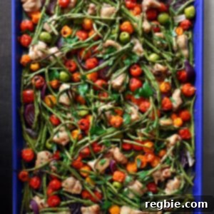 An overhead shot of a blue sheet pan filled with cooked cubed chicken thighs, roasted green beans, roasted cherry tomatoes, red onion wedges, green olives, fresh herbs, and a drizzle of balsamic vinegar.