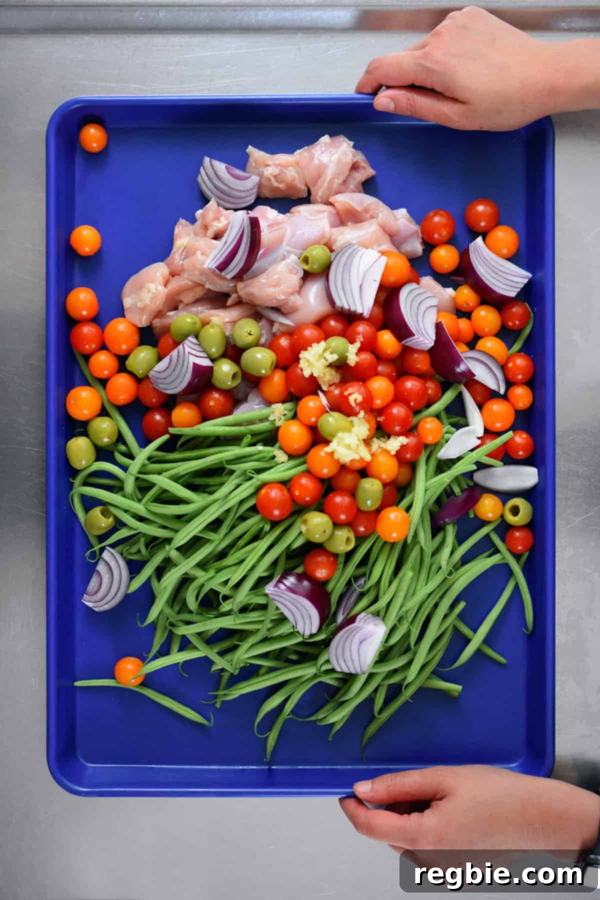 An overhead shot of the raw and unseasoned ingredients for sheet pan Italian chicken — cubed chicken thighs, green beans, cherry tomatoes, olives, and red onion — on a blue rimmed baking sheet, before cooking.