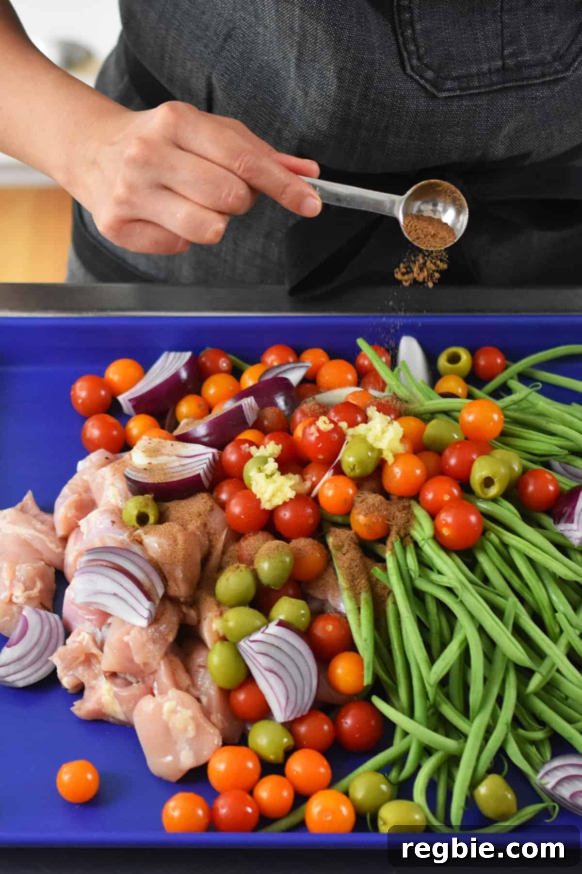 A hand adding a spoonful of Magic Mushroom Powder seasoning to a sheet pan filled with raw ingredients for Italian chicken, ensuring even distribution.