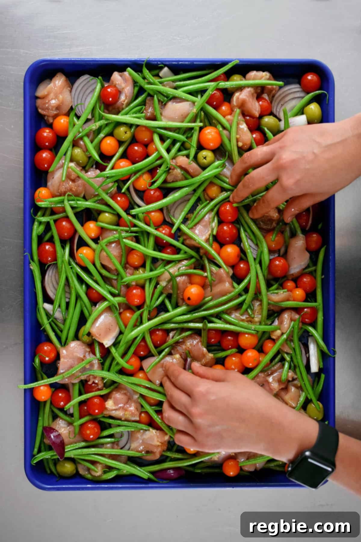 An overhead shot of two hands arranging the seasoned ingredients for sheet pan Italian chicken into a single, even layer on a blue rimmed baking sheet, ready for the oven.