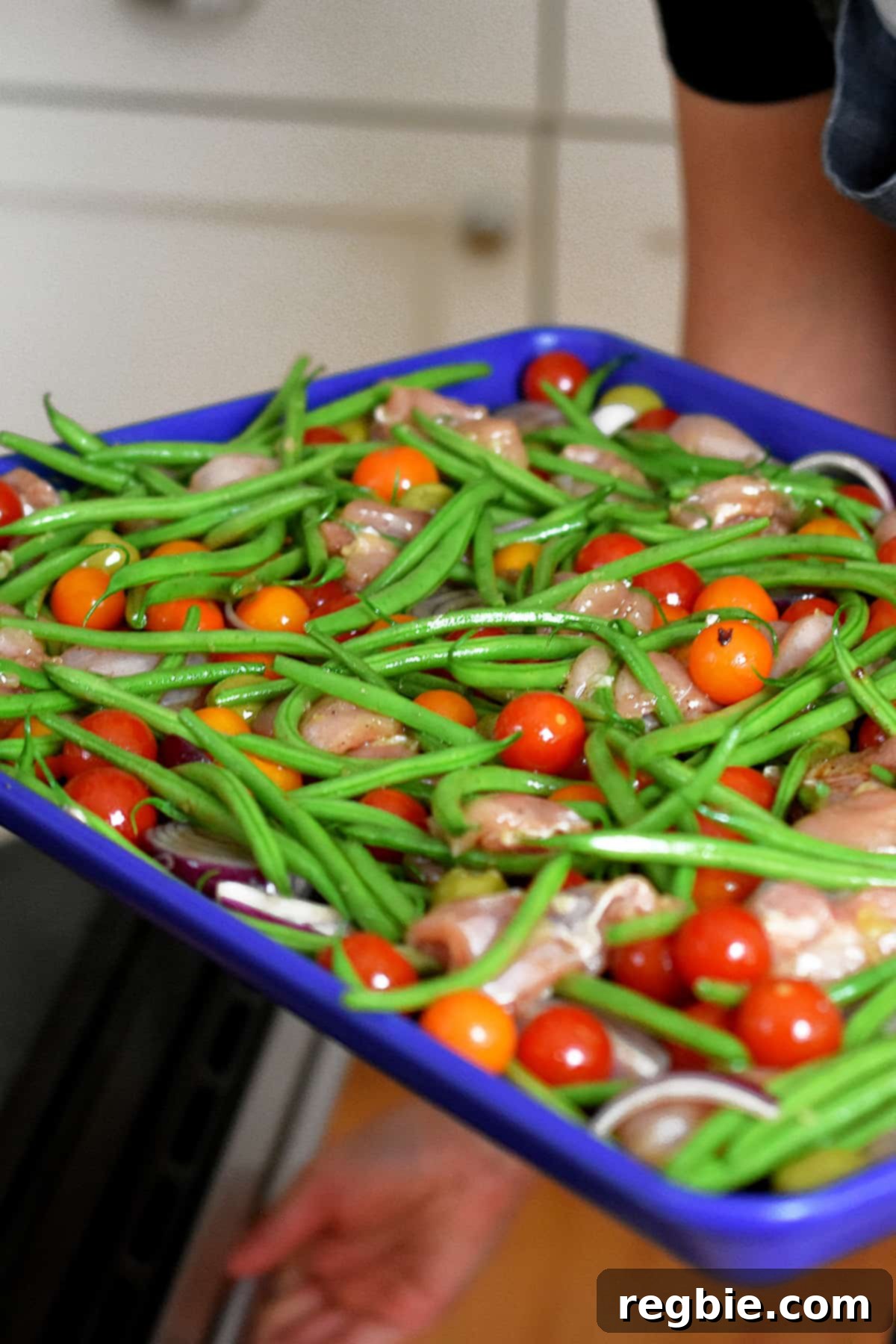 A person carrying a blue sheet pan filled with the seasoned ingredients for Italian chicken towards an oven, about to bake.