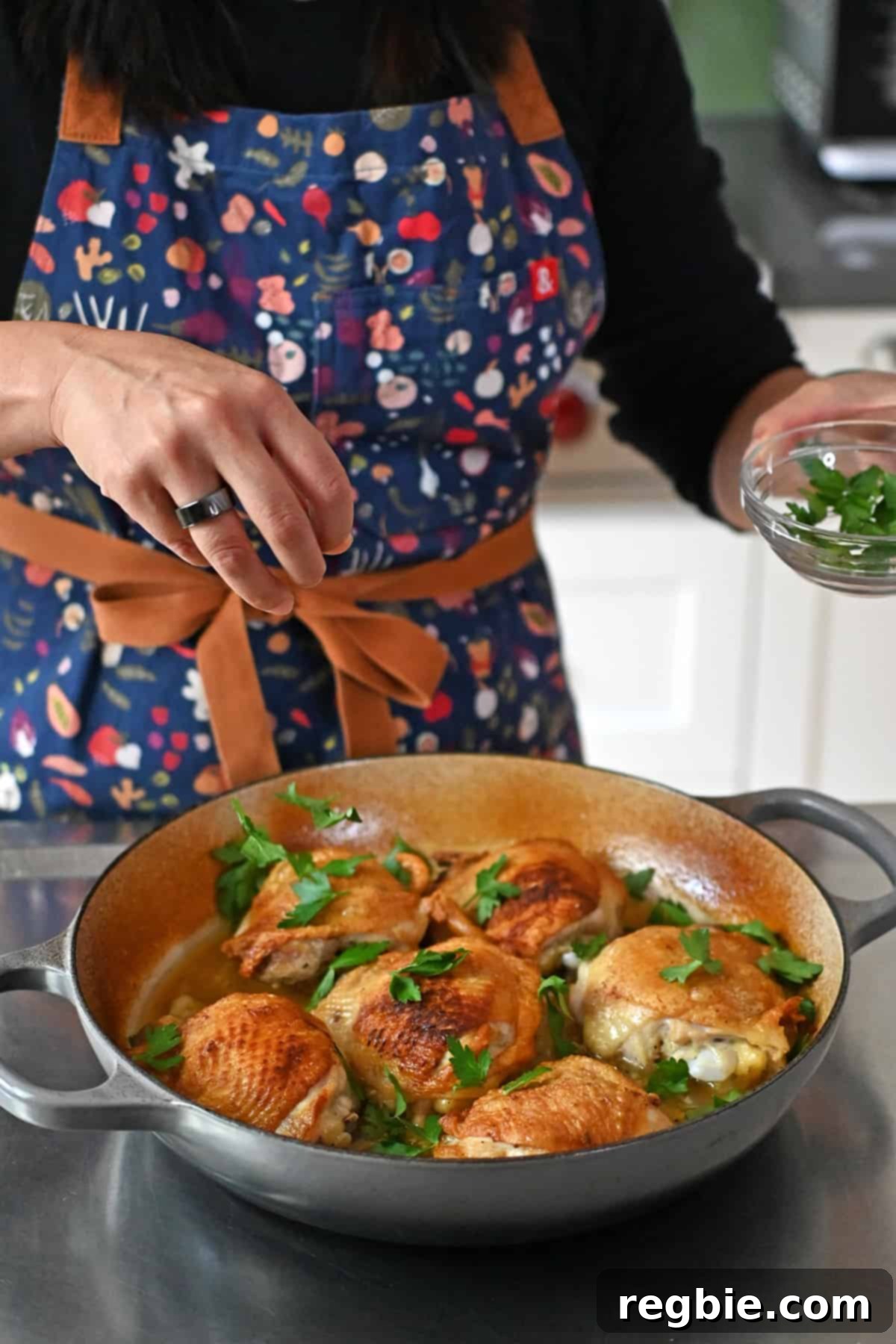A person in a blue apron is sprinkling fresh parsley leaves onto a skillet filled with lemon garlic chicken.