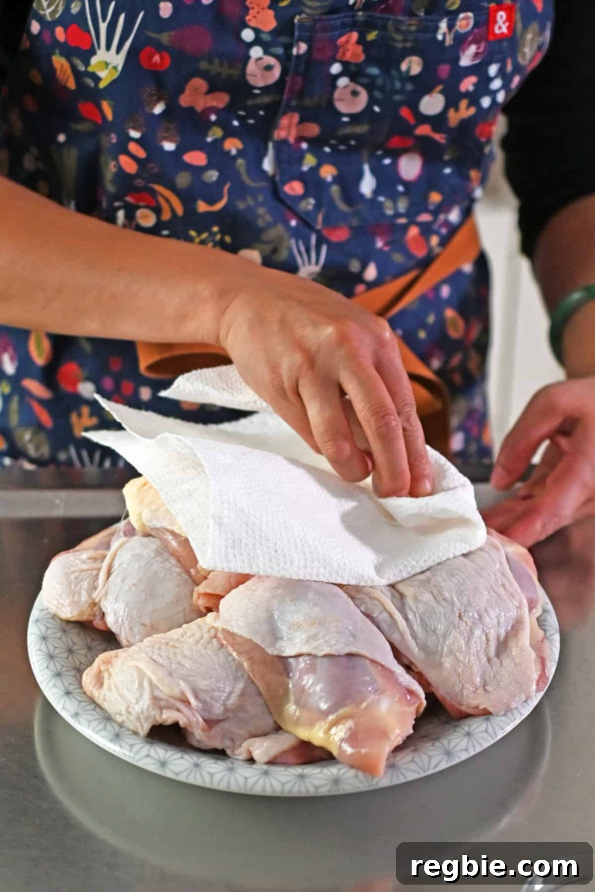 A person in a blue apron is blotting raw skin-on bone-in chicken thighs dry with a paper towel.