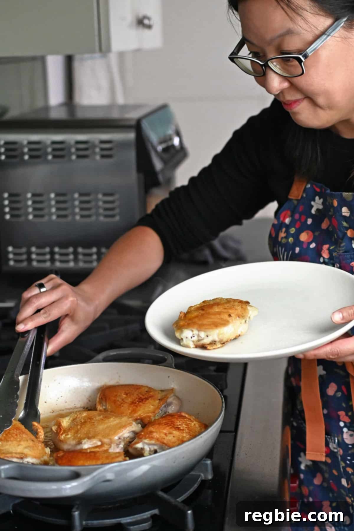 An Asian woman in glasses is transferring chicken thighs with golden brown skin from a skillet onto a plate with a pair of tongs.