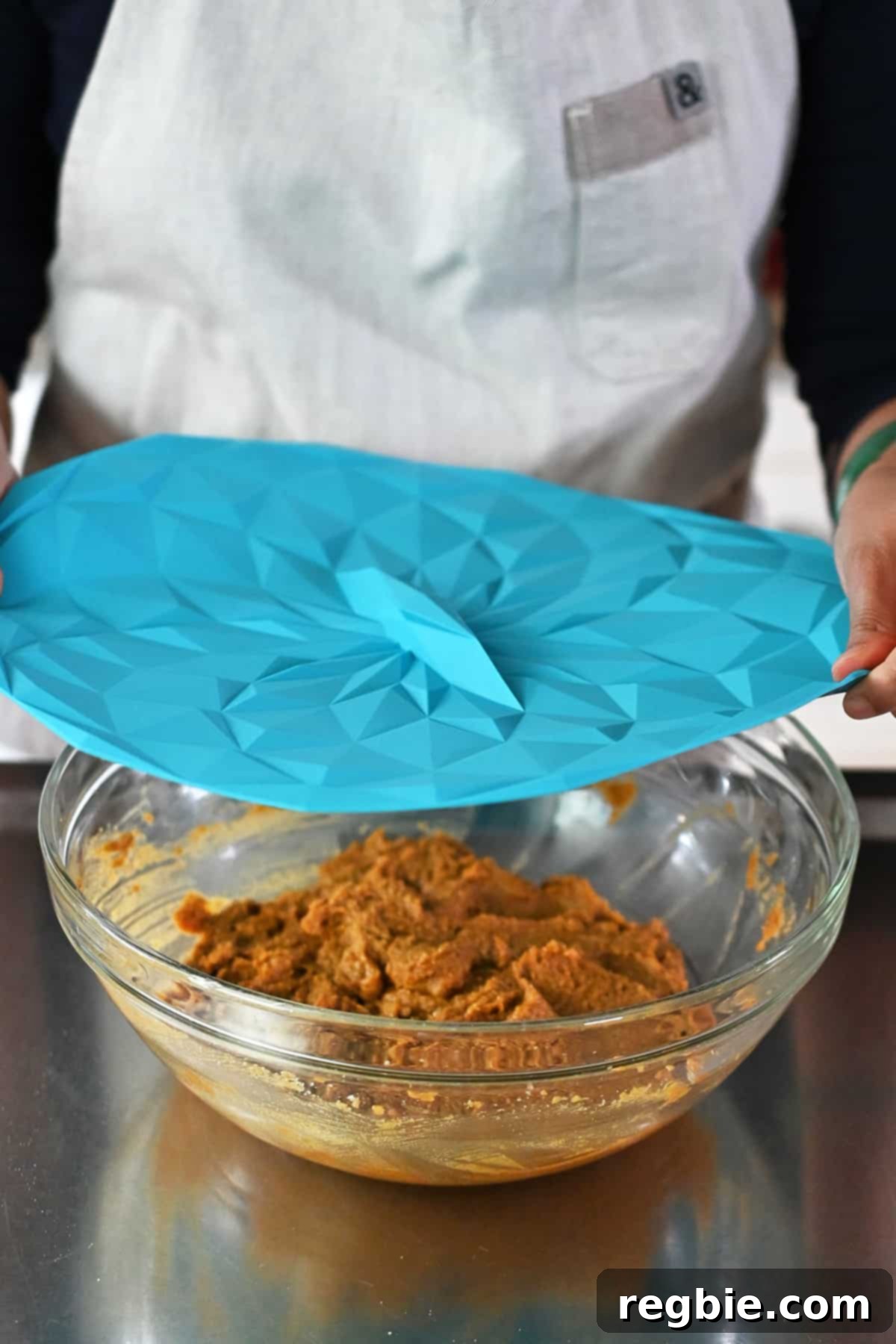 Paleo Pumpkin Bliss Cookies 14 A person is placing a blue silicone lid on top of a bowl filled with uncooked paleo pumpkin cookie dough before chilling it in the fridge.