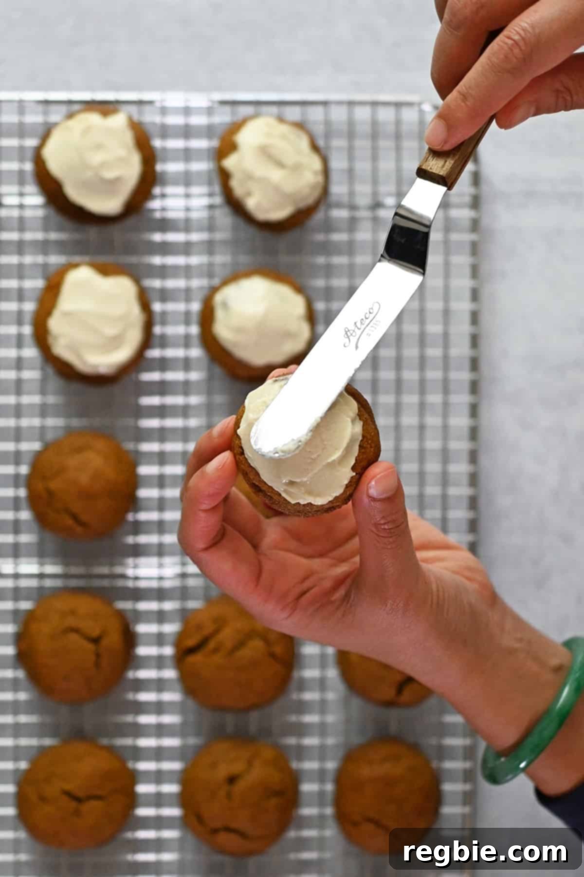 Paleo Pumpkin Bliss Cookies 19 A hand is holding a paleo pumpkin cookie and spreading dairy-free cream cheese frosting on top with a small offset spatula.