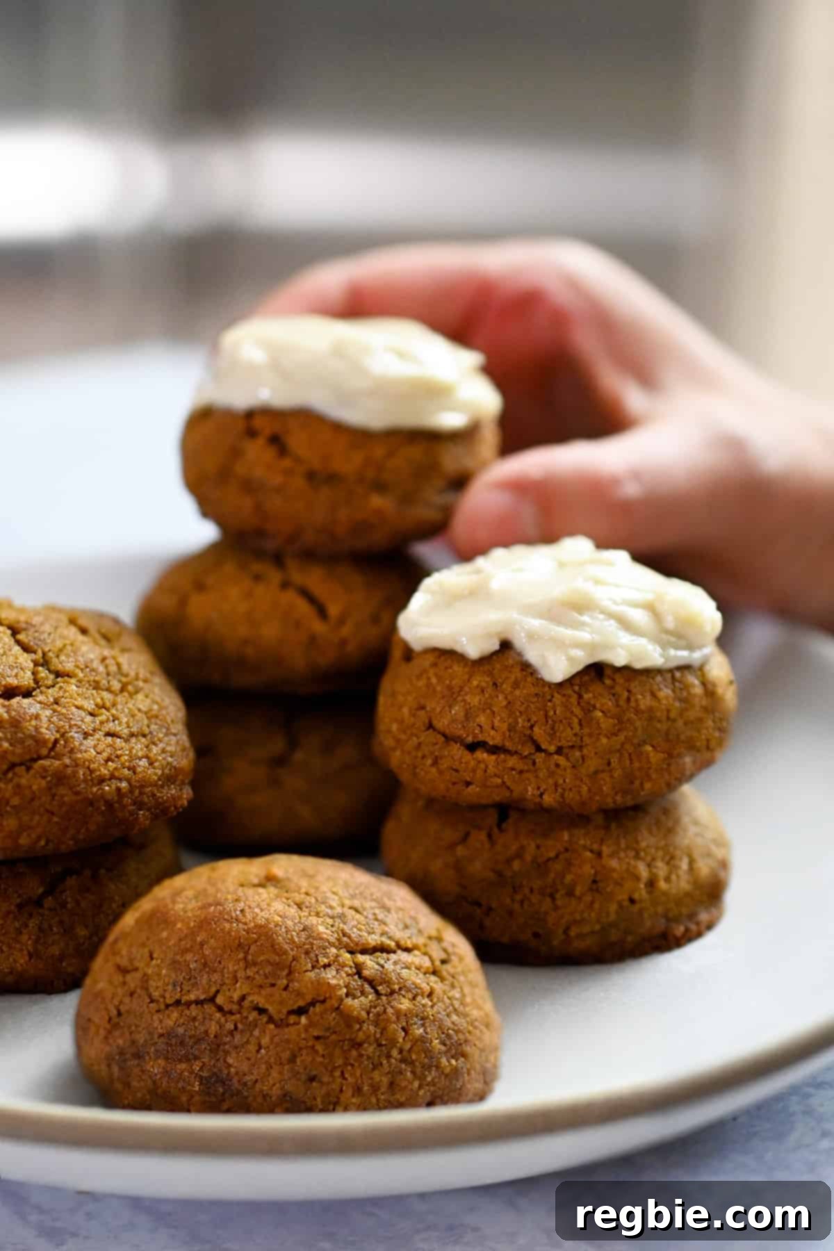 Paleo Pumpkin Bliss Cookies 3 A hand is grabbing a dairy-free cream-cheese frosted pumpkin cookie from a plate filled with cookies.