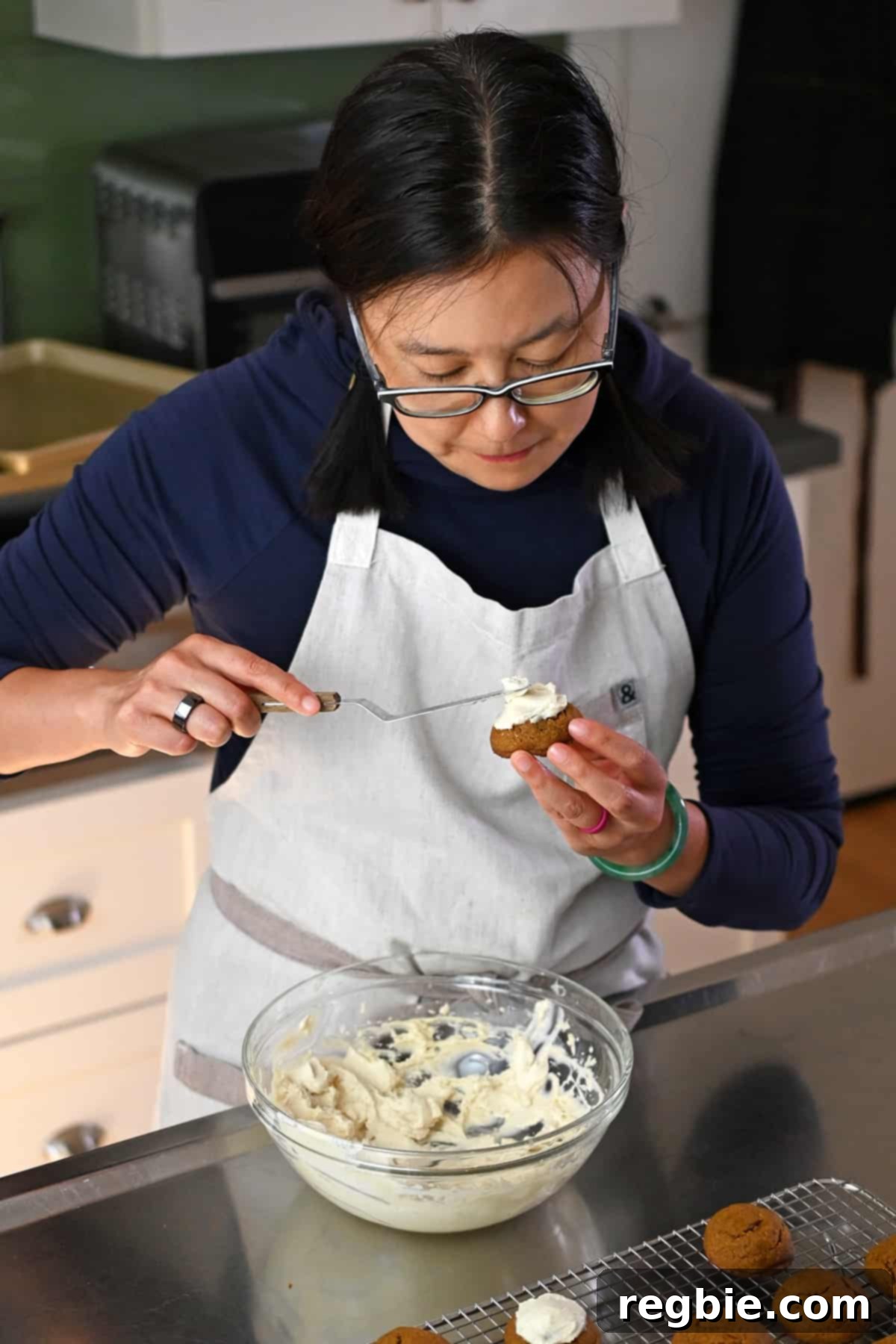 Paleo Pumpkin Bliss Cookies 5 An Asian woman in glasses is using a small offset spatula to add dairy-free cream cheese frosting onto paleo pumpkin cookies.