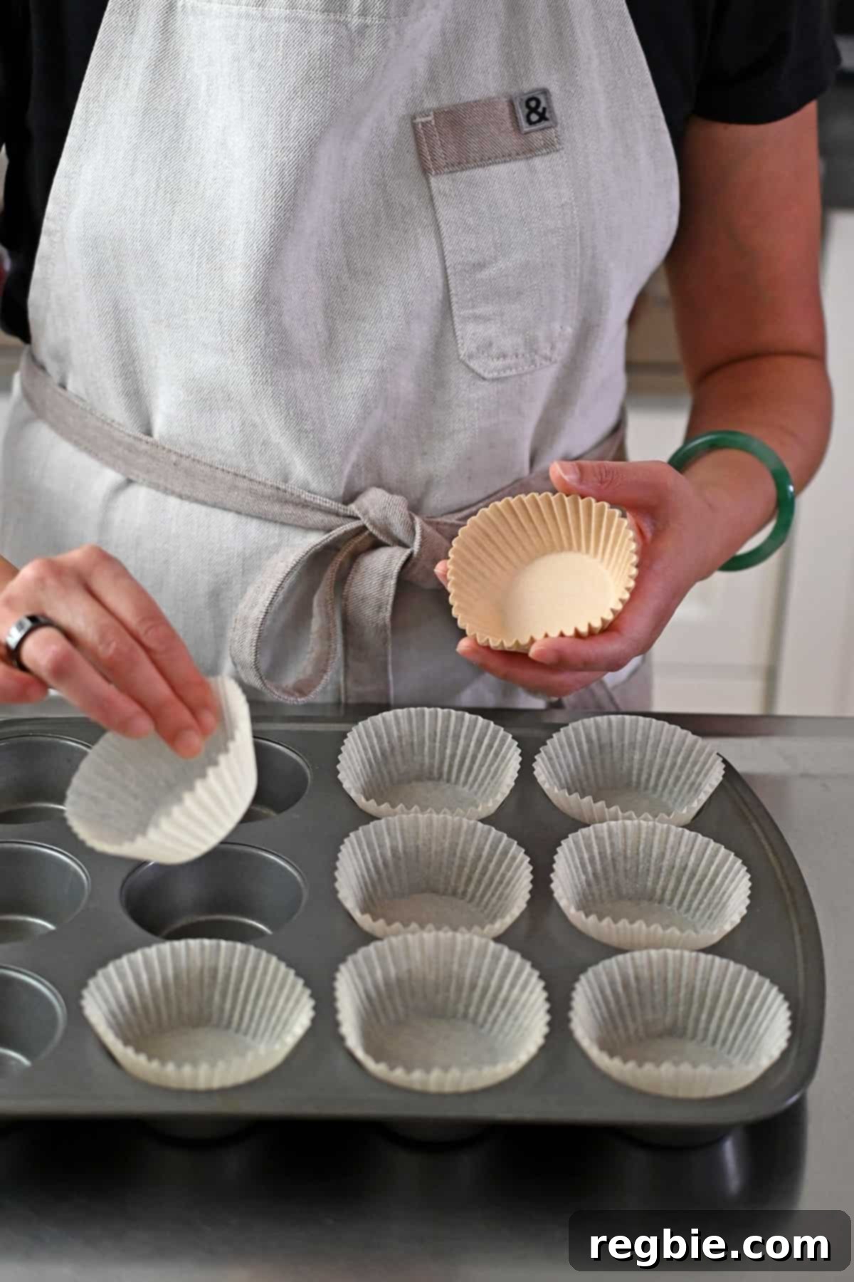A person in a beige apron is placing parchment liners into a muffin tin, ready for batter.