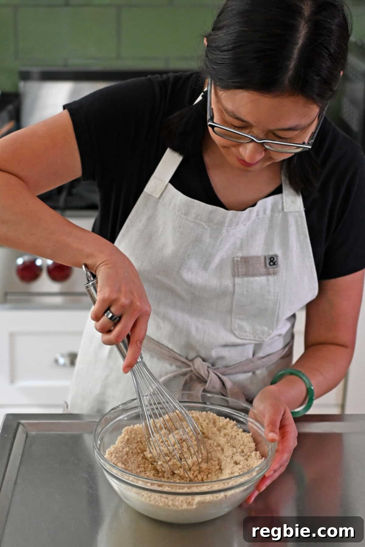 An Asian woman in glasses is mixing the dry ingredients for paleo apple muffins in a clear glass mixing bowl with a metal whisk.