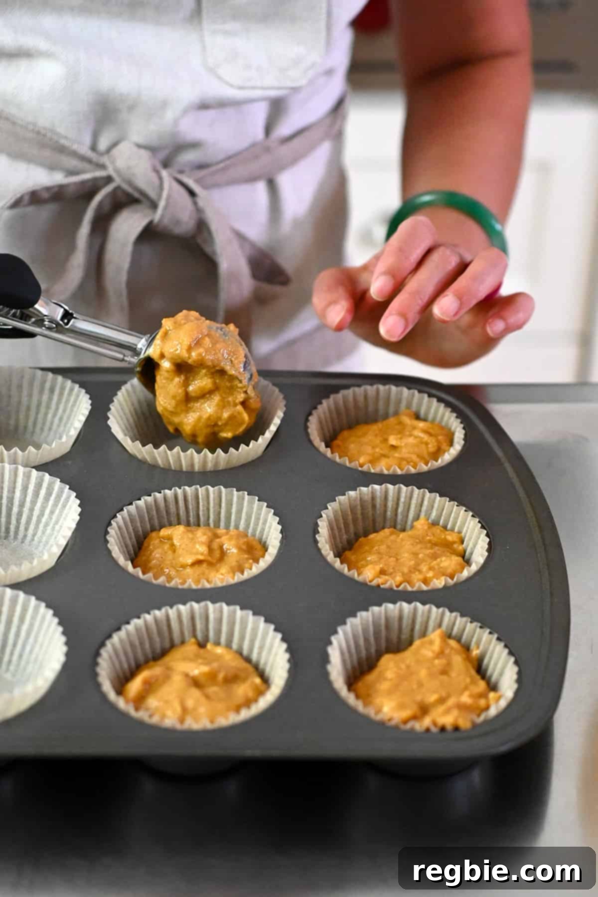 Adding the apple muffin batter with an ice cream scoop to parchment muffin liners in a muffin tin.