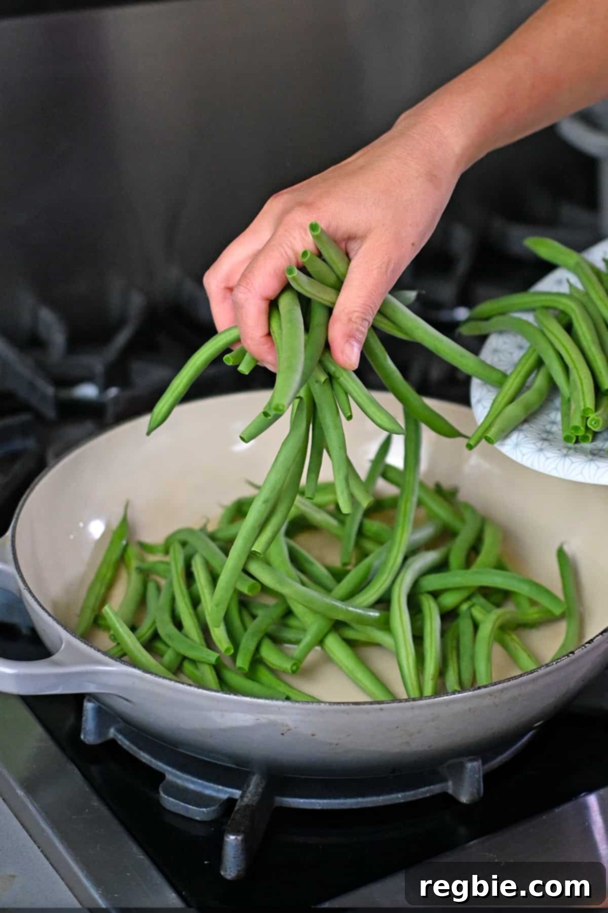 Adding fresh, trimmed green beans to an enamel cast iron skillet with hot oil for stir-frying.