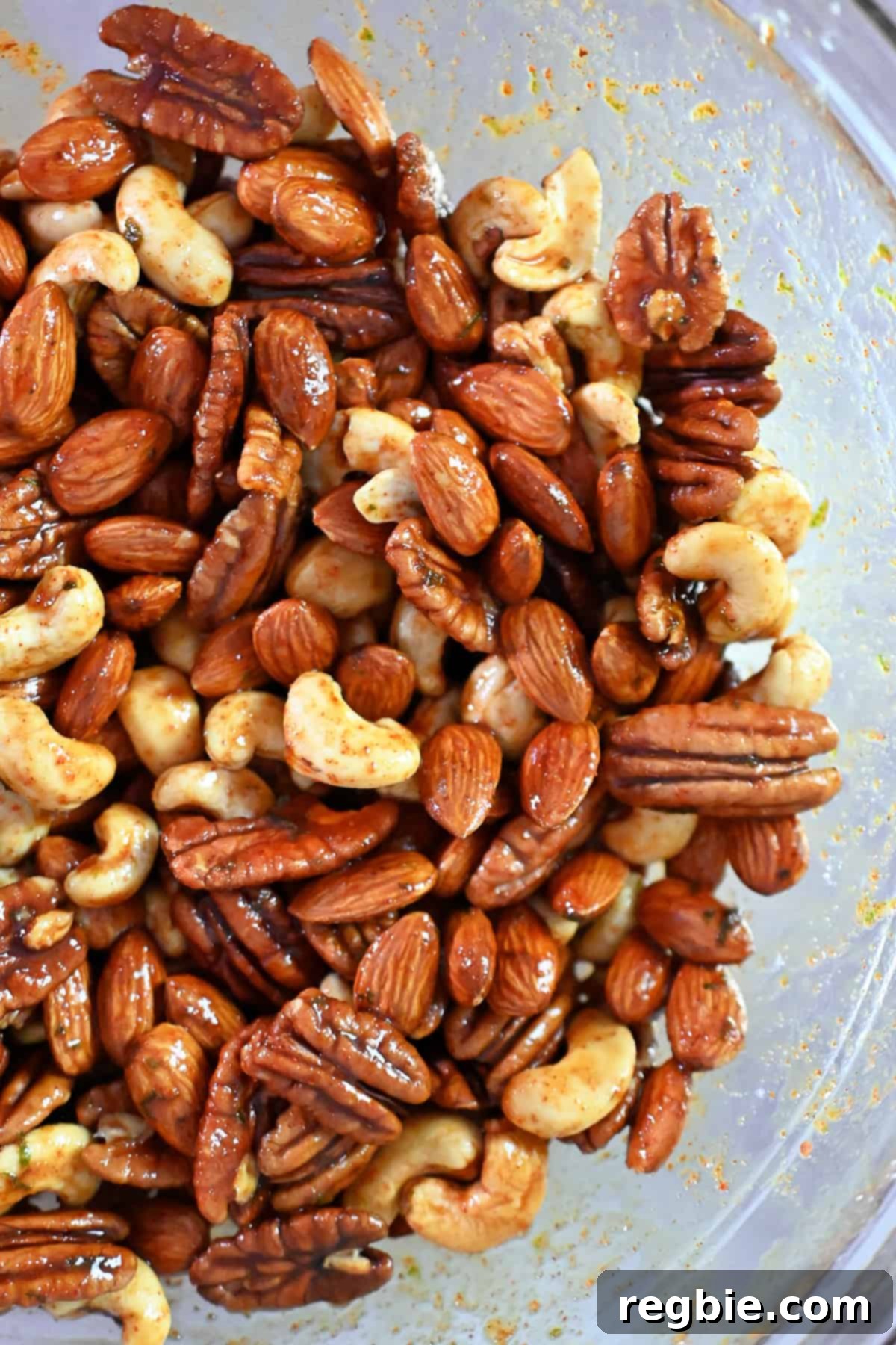 A close-up of raw mixed nuts being perfectly tossed in a rich, spice-packed sauce within a large glass bowl, ensuring every nut is coated.