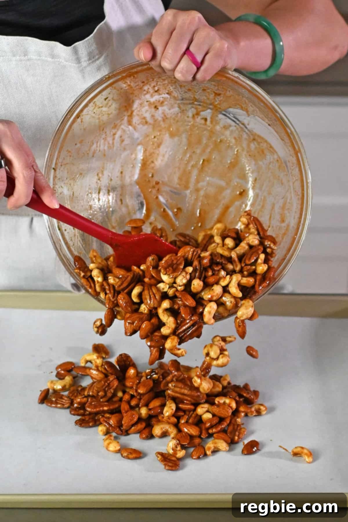 The process of transferring the perfectly seasoned nuts from a large glass mixing bowl onto a parchment-lined, rimmed baking sheet.