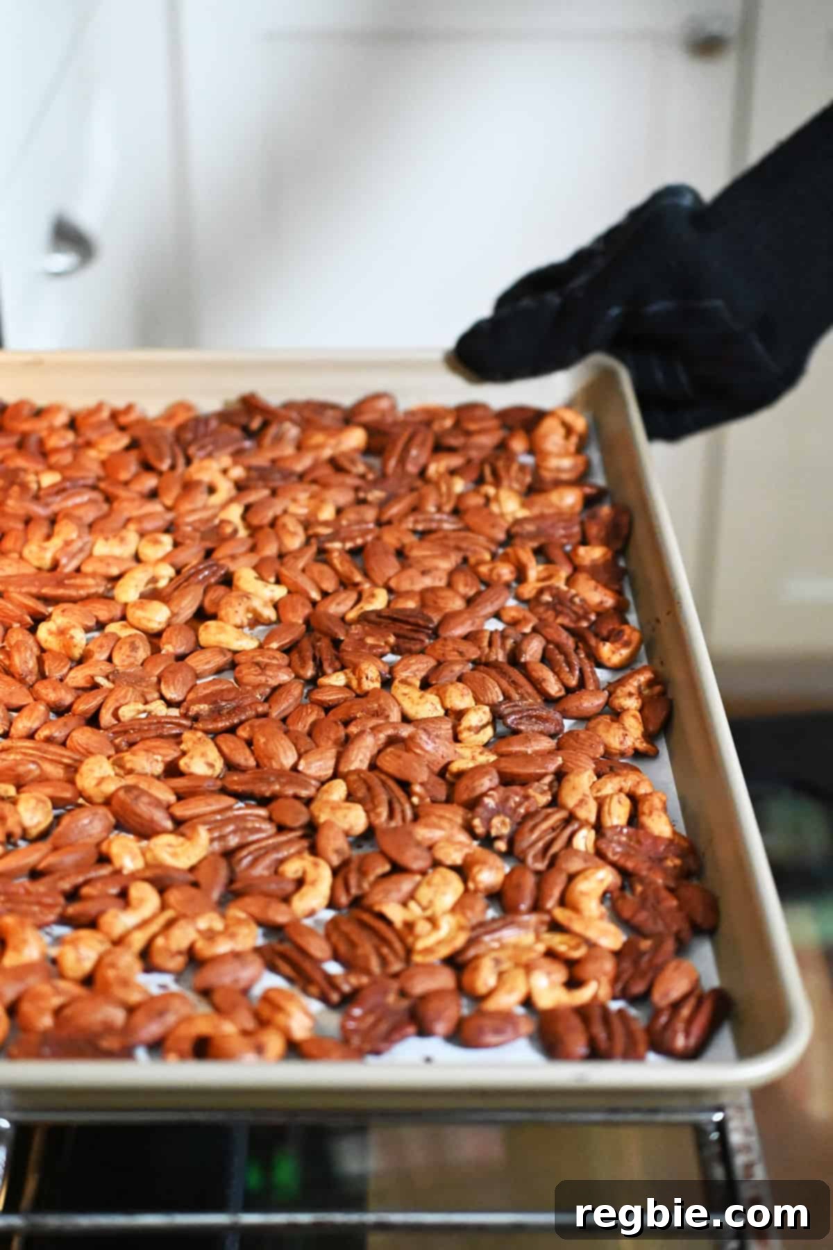 A hand carefully removing a rimmed baking sheet filled with freshly roasted, perfectly golden spice nuts from an open oven.
