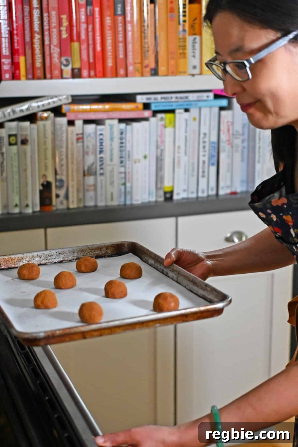 A smiling Asian woman carefully placing a tray of paleo and gluten-free snickerdoodle cookie dough balls into the preheated oven.