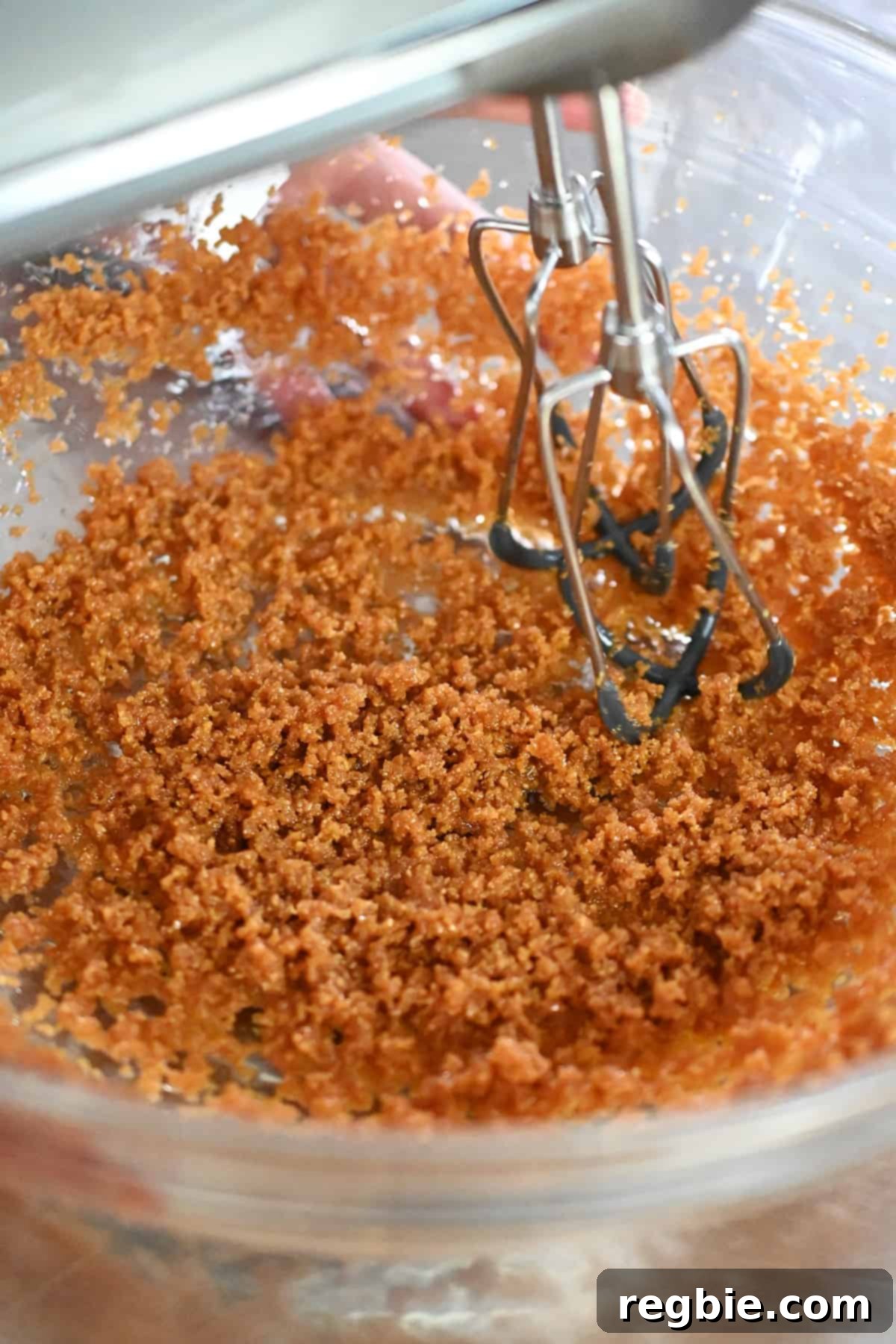 A close-up view of the blended coconut sugar, coconut oil, and vanilla extract in a glass mixing bowl, showing the consistency of wet sand.