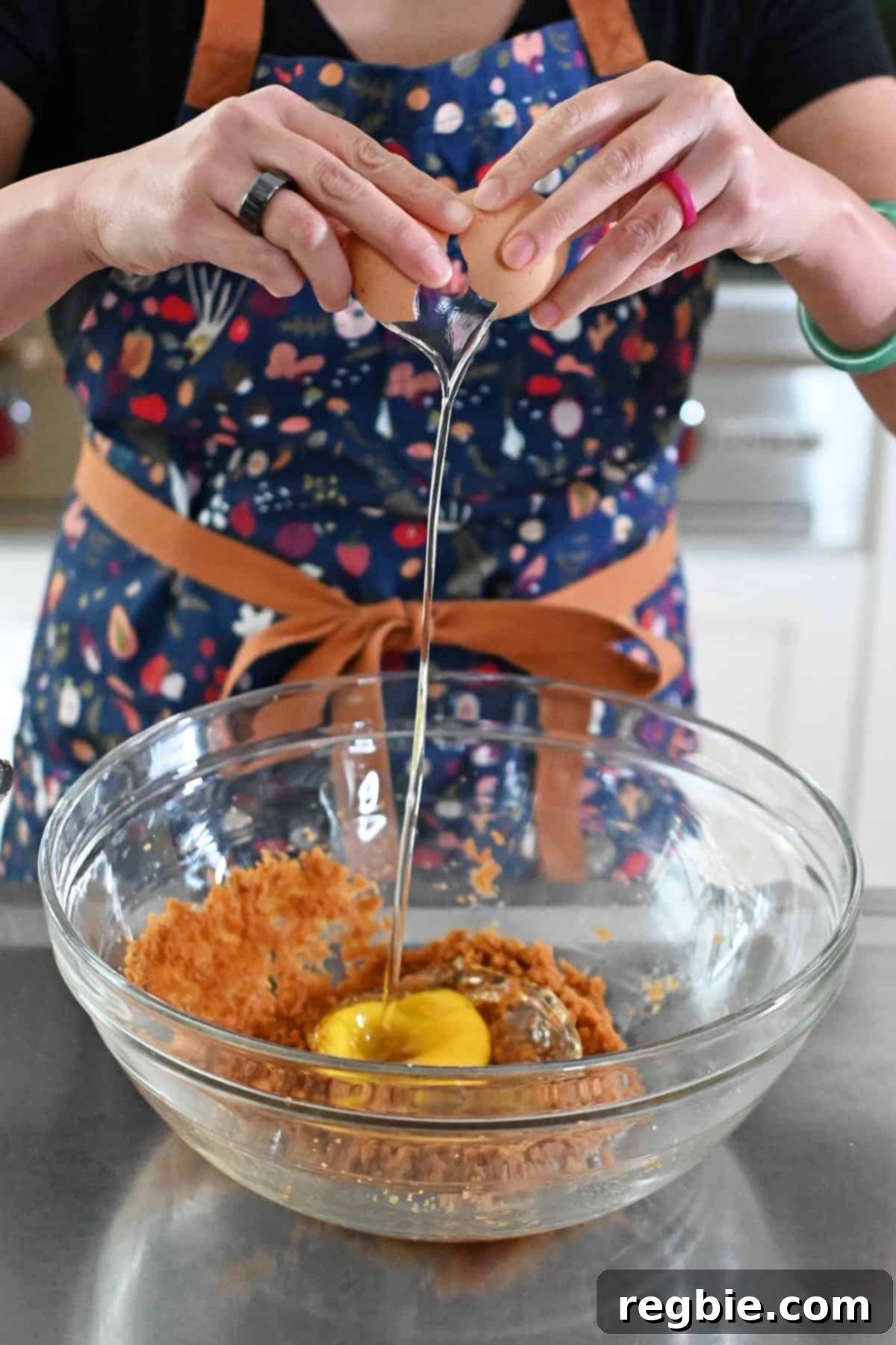 A person in a blue apron cracking an egg into a bowl already containing coconut sugar, melted coconut oil, and vanilla extract.