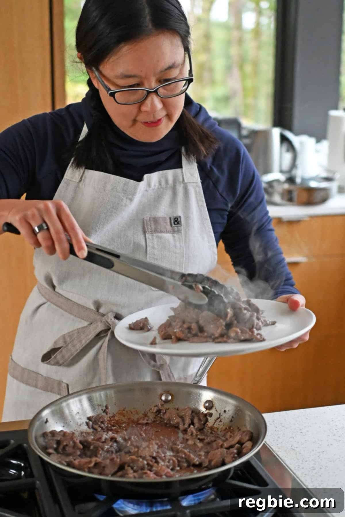 An Asian woman transfers cooked sliced flank steak from a skillet to a plate with a pair of tongs.