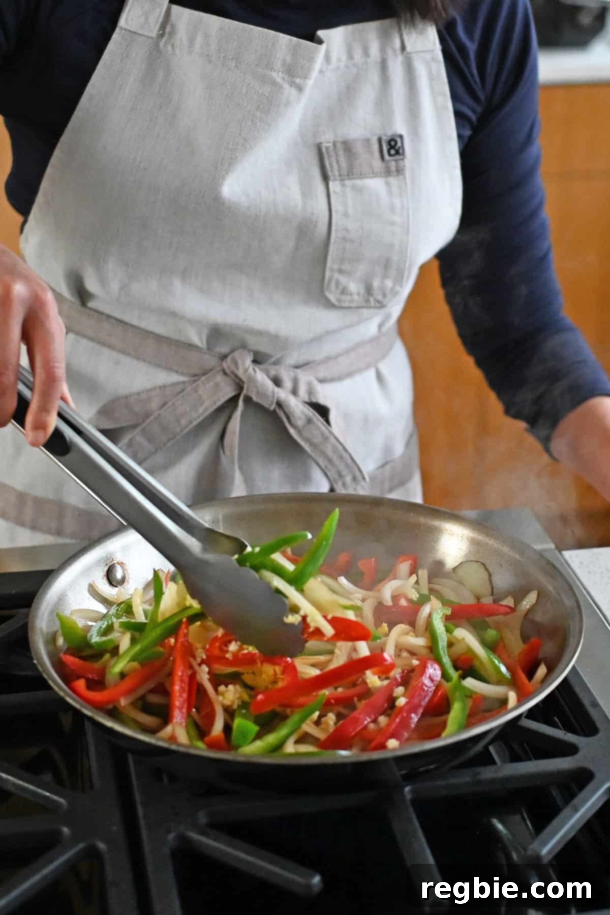 A pair of tongs is tossing sliced onions and belle peppers in a stainless steel skillet.