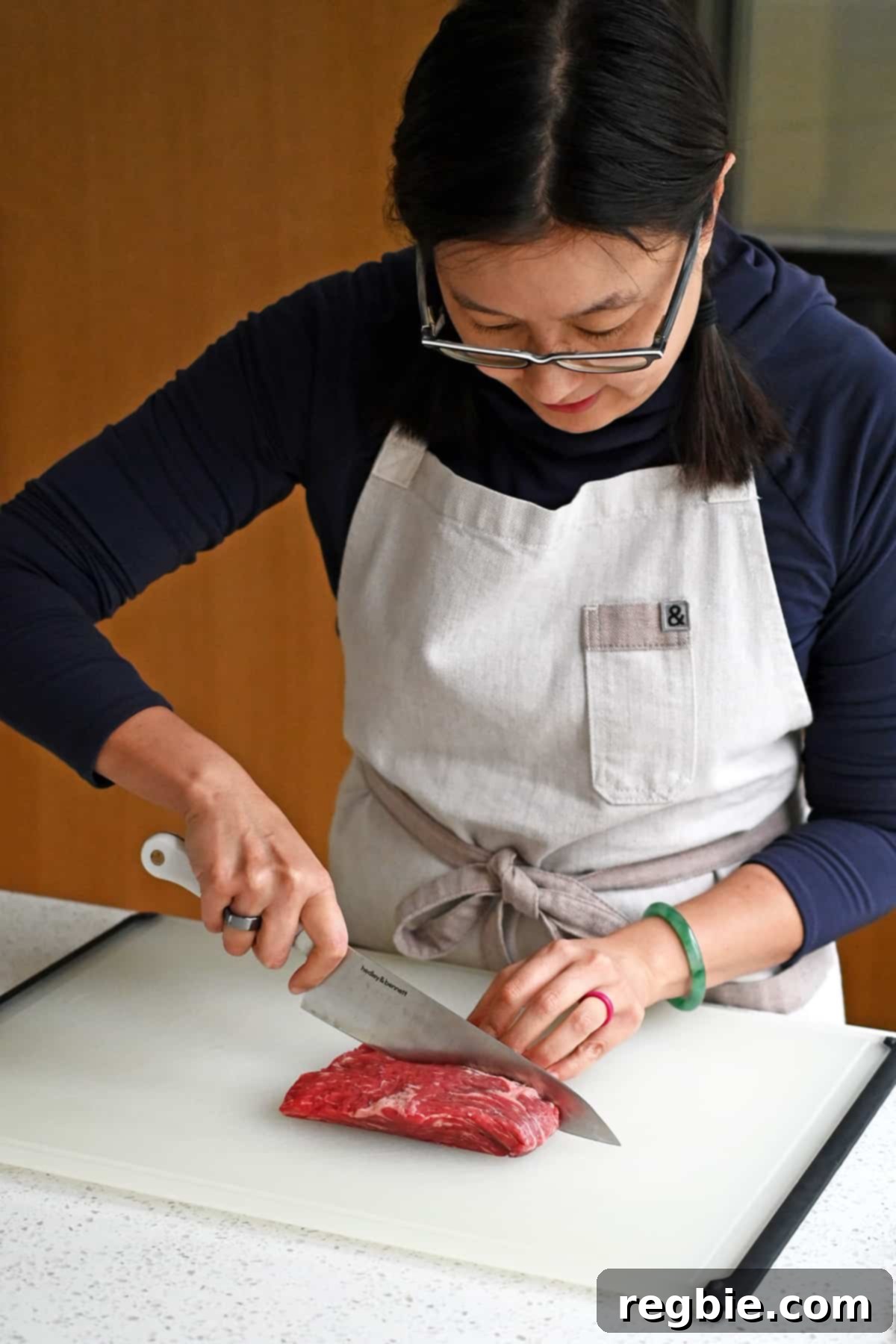 An Asian woman is slicing a raw piece of flank steakin half with the grain.