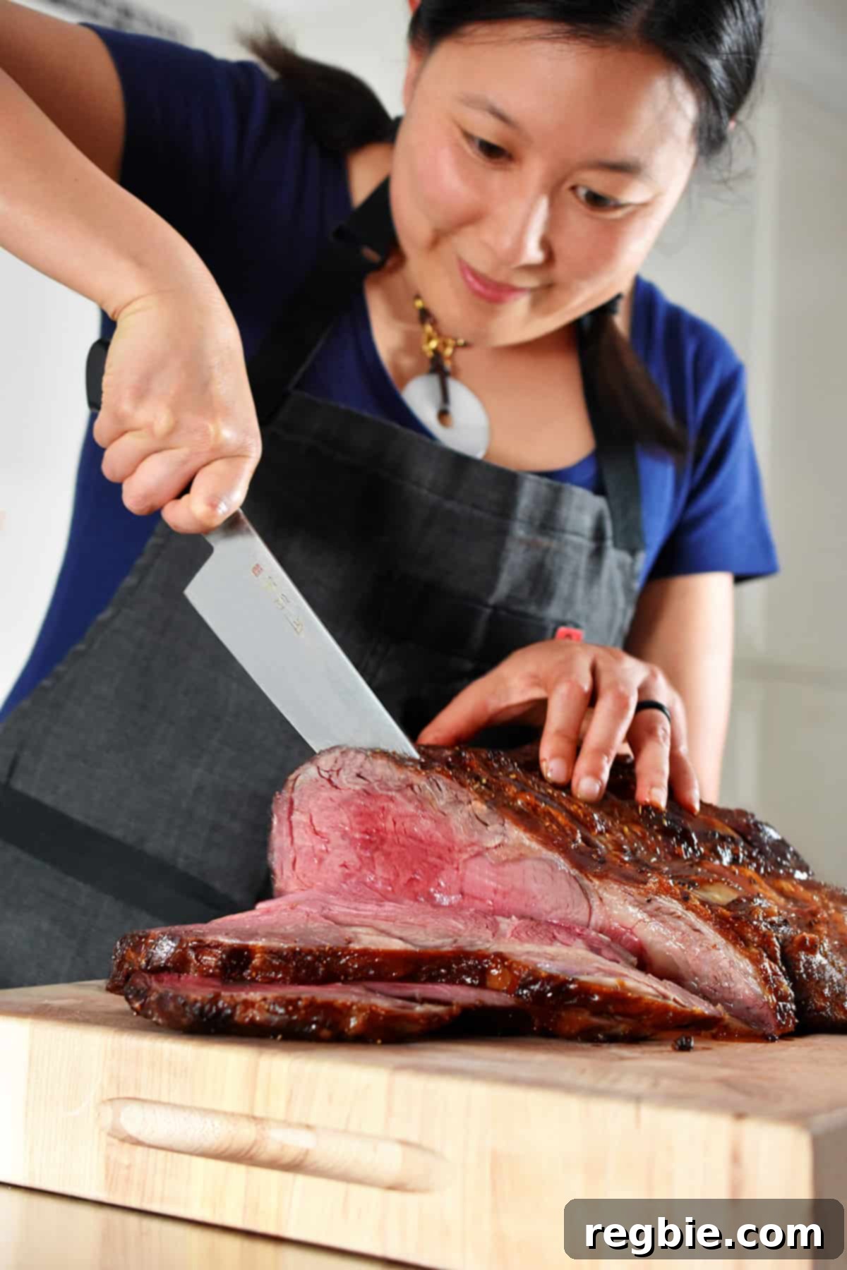 A smiling Asian woman expertly slicing a medium-rare prime rib roast into thin, even slices on a sturdy wooden cutting board.