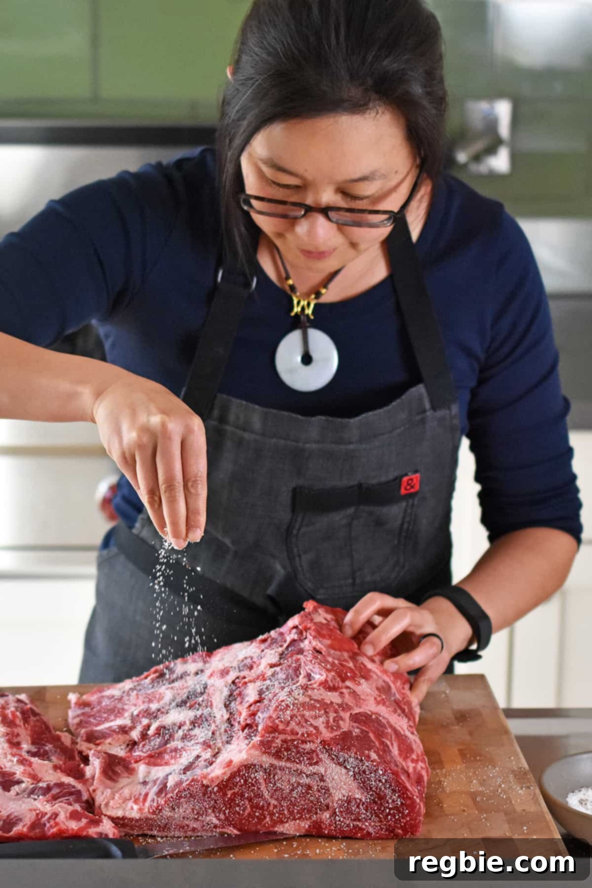An Asian woman with glasses sprinkling a generous amount of salt and pepper onto a boneless prime rib roast, ensuring even coverage.