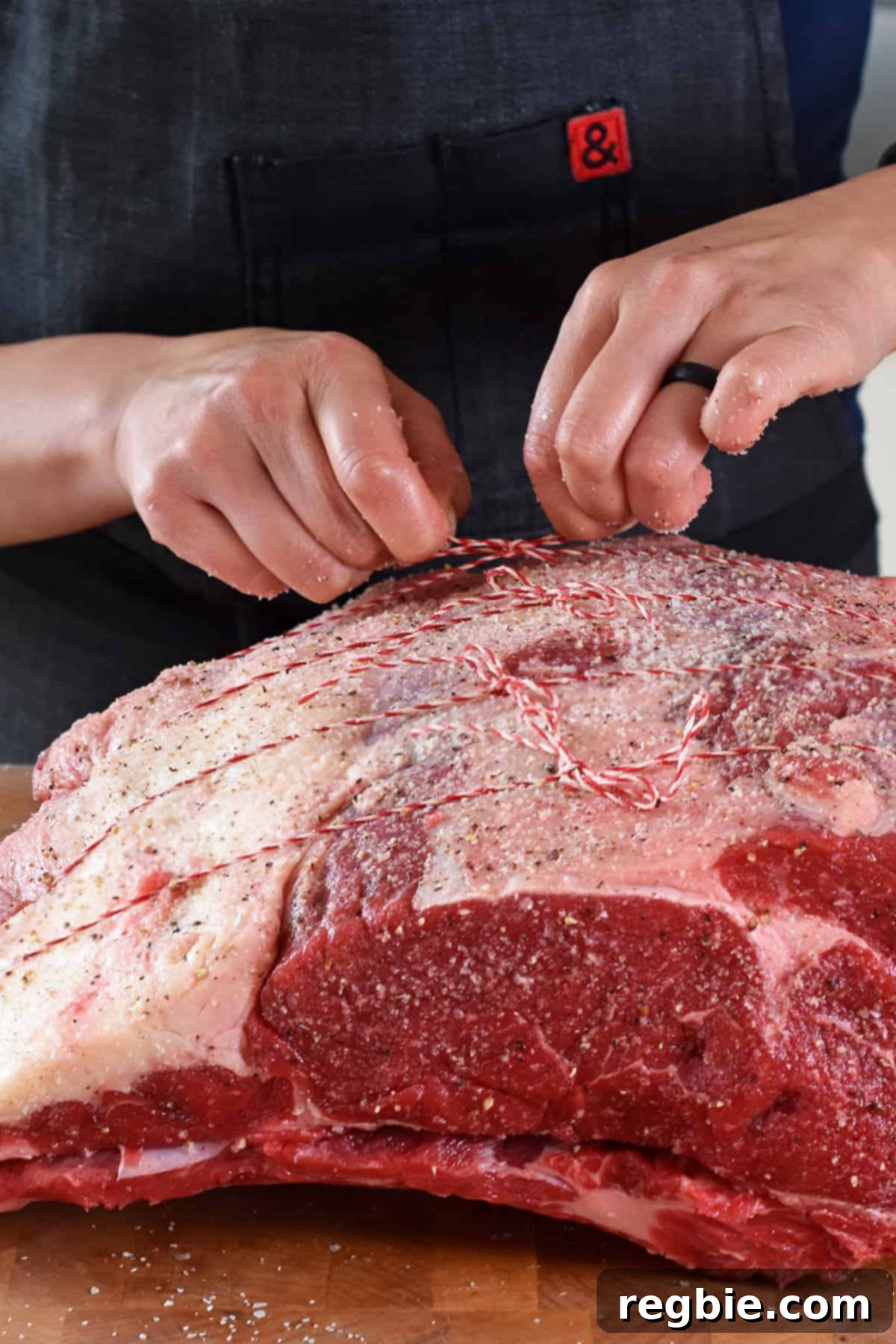 Hands carefully tying a seasoned boneless prime rib roast onto the detached bone rack with kitchen twine, ready for dry-brining.